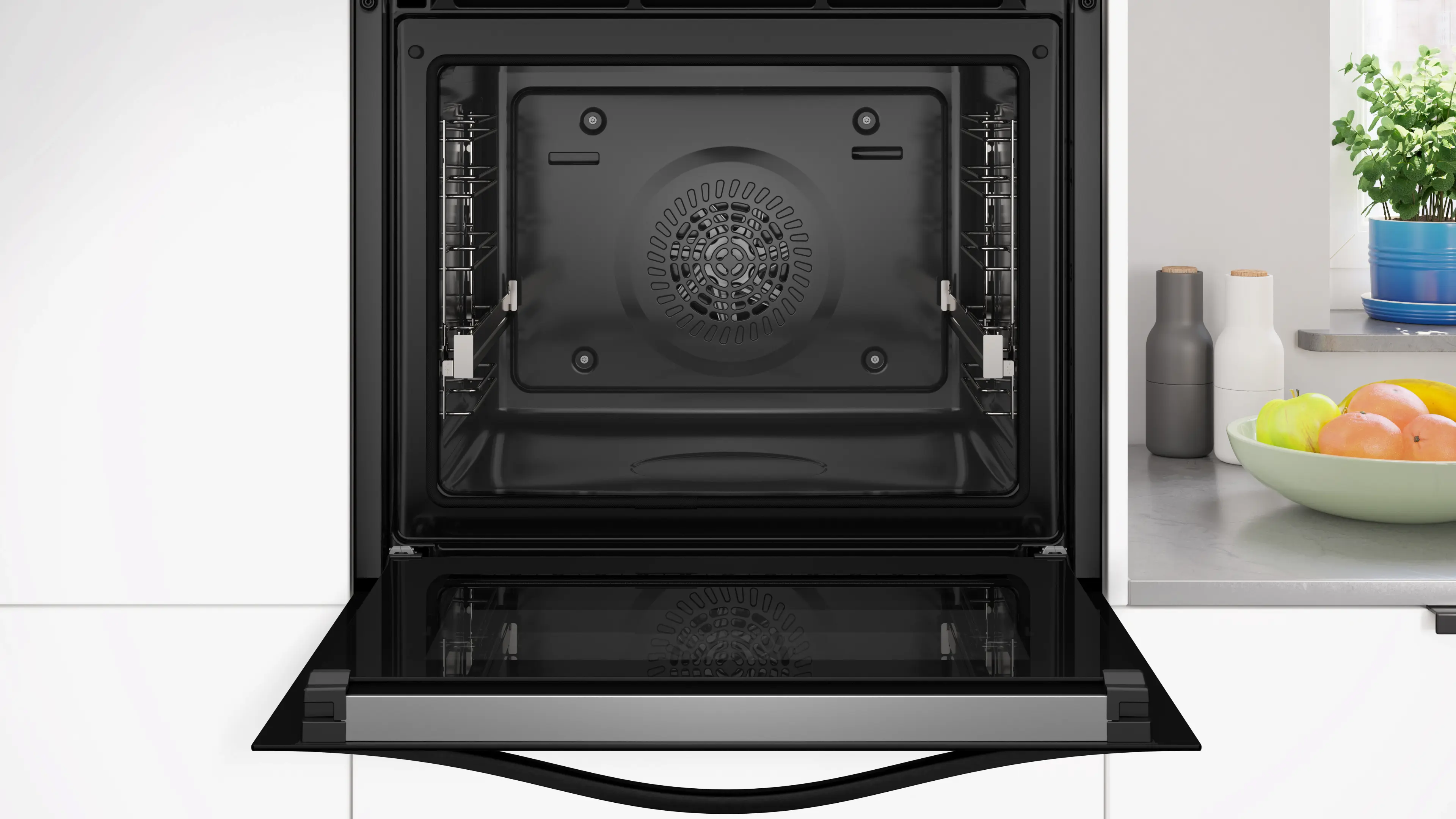 Horno abierto con interior negro. Dentro, se ve un ventilador. Encimera de cocina con frutas y plantas a la derecha.