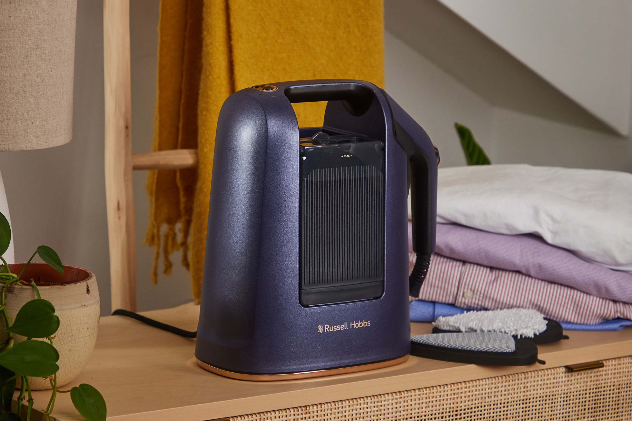 Egy kék Russell Hobbs gőzölő fa felületen. Mögötte sárga takaró és hajtogatott ruhák láthatók.