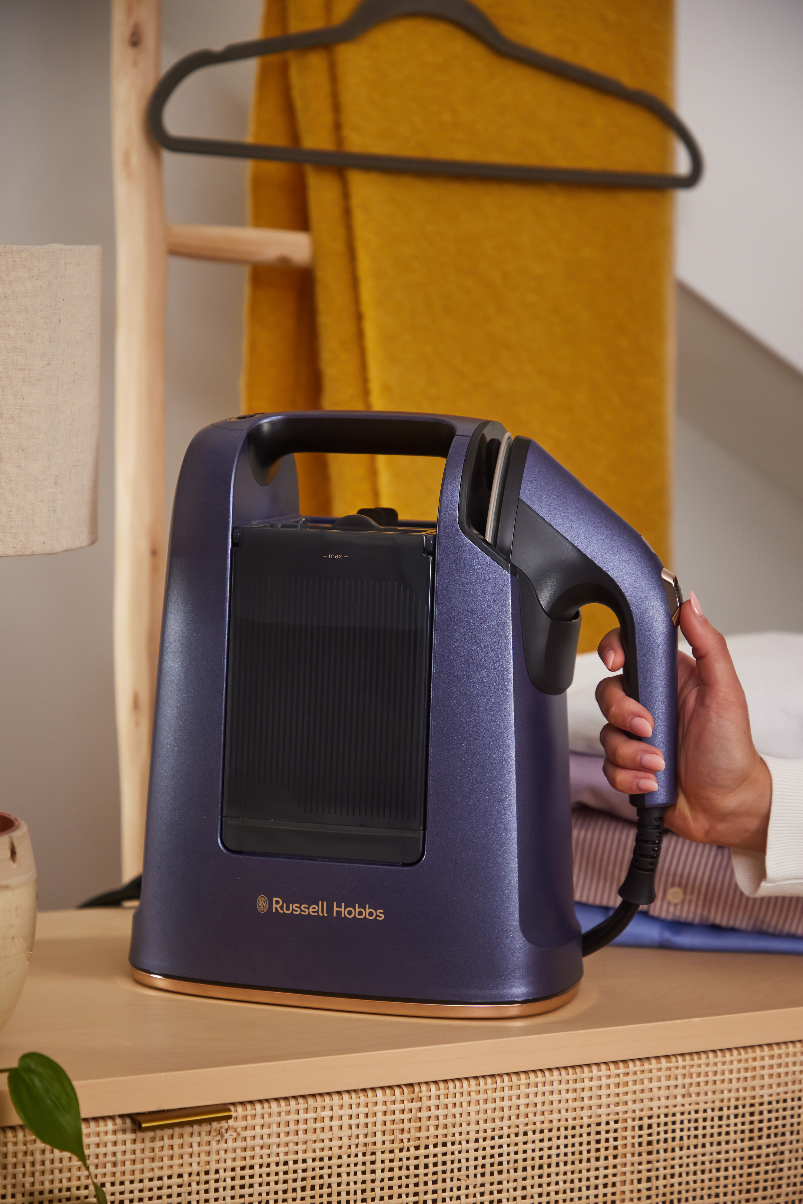 Egy lila Russell Hobbs gőzölős vasaló egy asztalon. Egy kéz fogja a fogantyút. Sárga takaró a háttérben.
