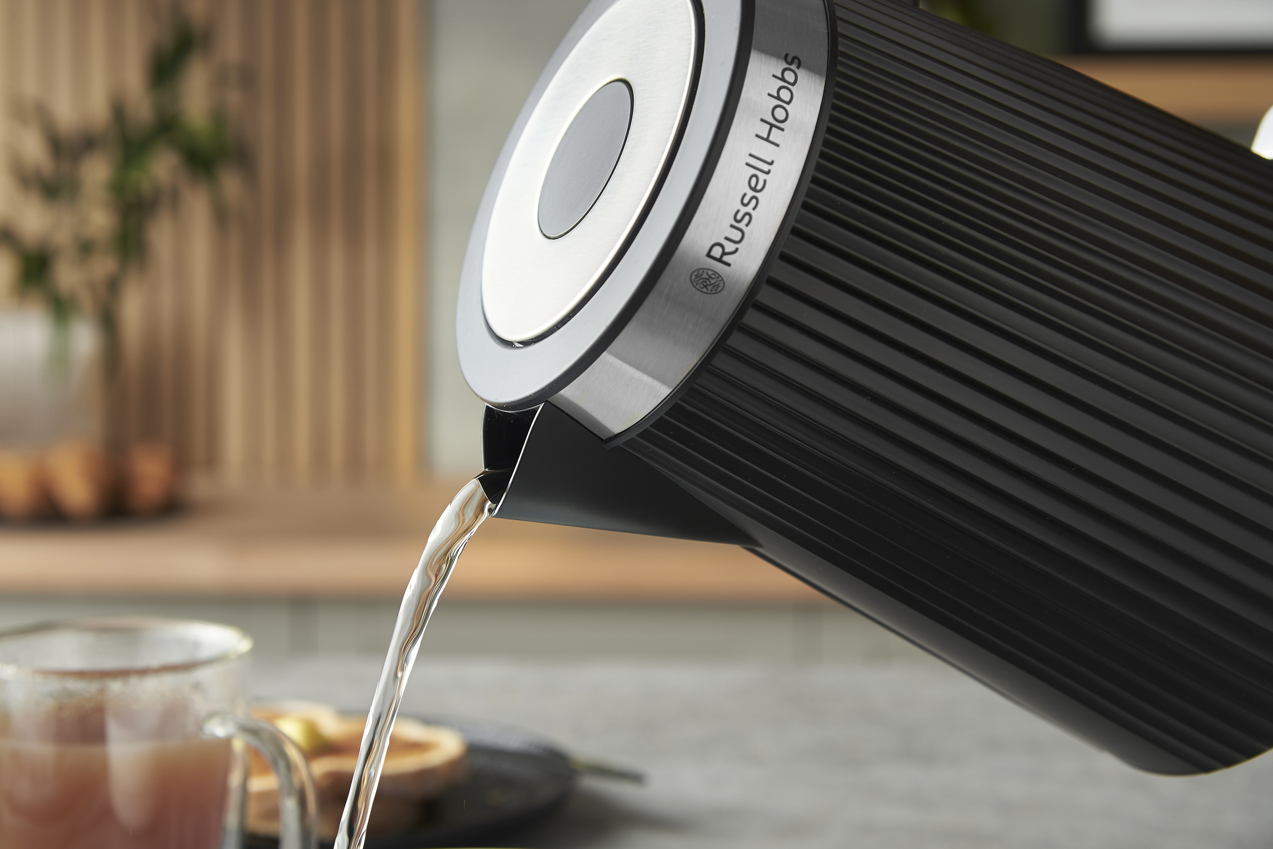 Egy fekete Russell Hobbs vízforraló vizet önt egy pohárba. A háttérben egy elmosódott konyhai jelenet látható.