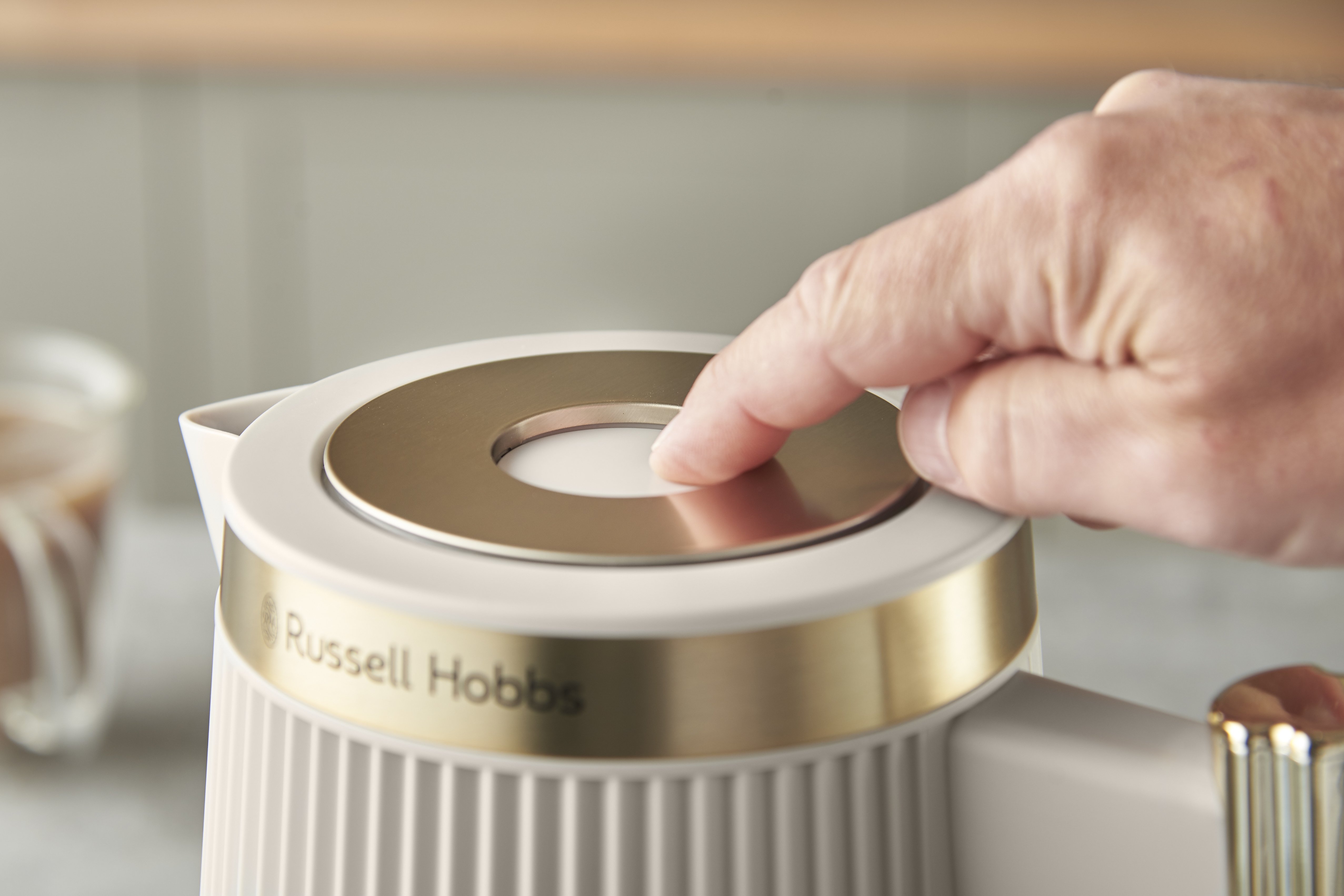 Egy kéz megnyomja a Russell Hobbs vízforraló gombját. A vízforraló törtfehér, arany díszítéssel. Homályos háttér.