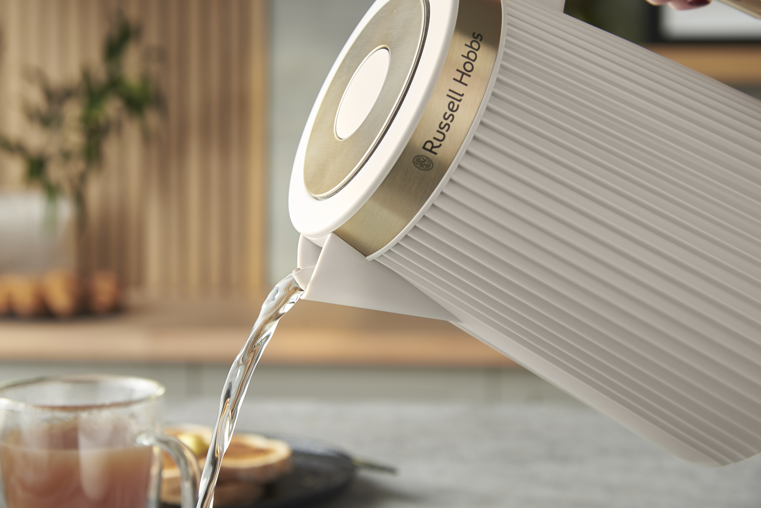Egy fehér Russell Hobbs vízforraló vizet önt egy pohárba. A háttérben pirítós és egy csésze látható.