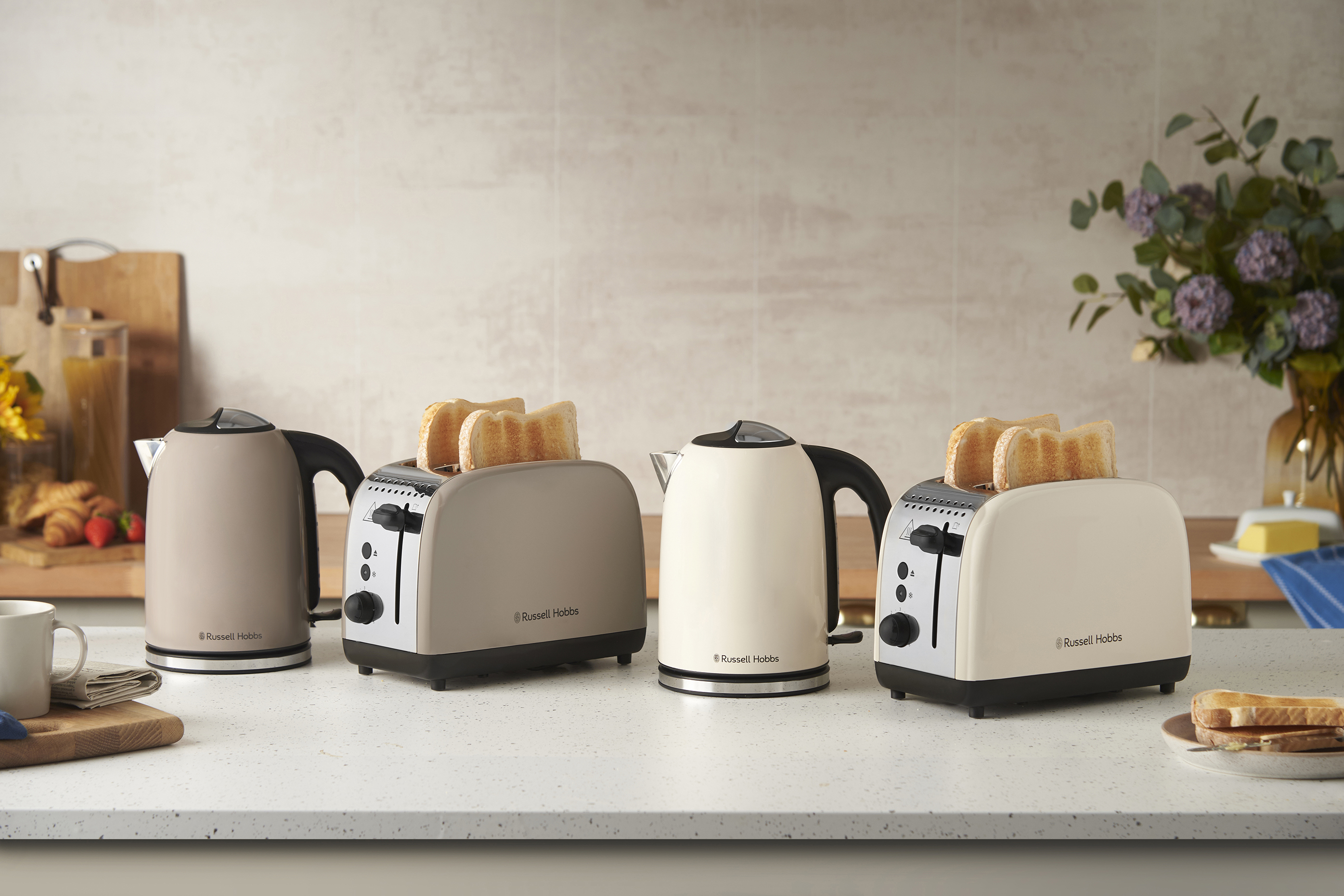 Négy Russell Hobbs készülék a pulton: két kenyérpirító pirítóssal, és két vízforraló. A háttérben virágok, vágódeszka és vaj.