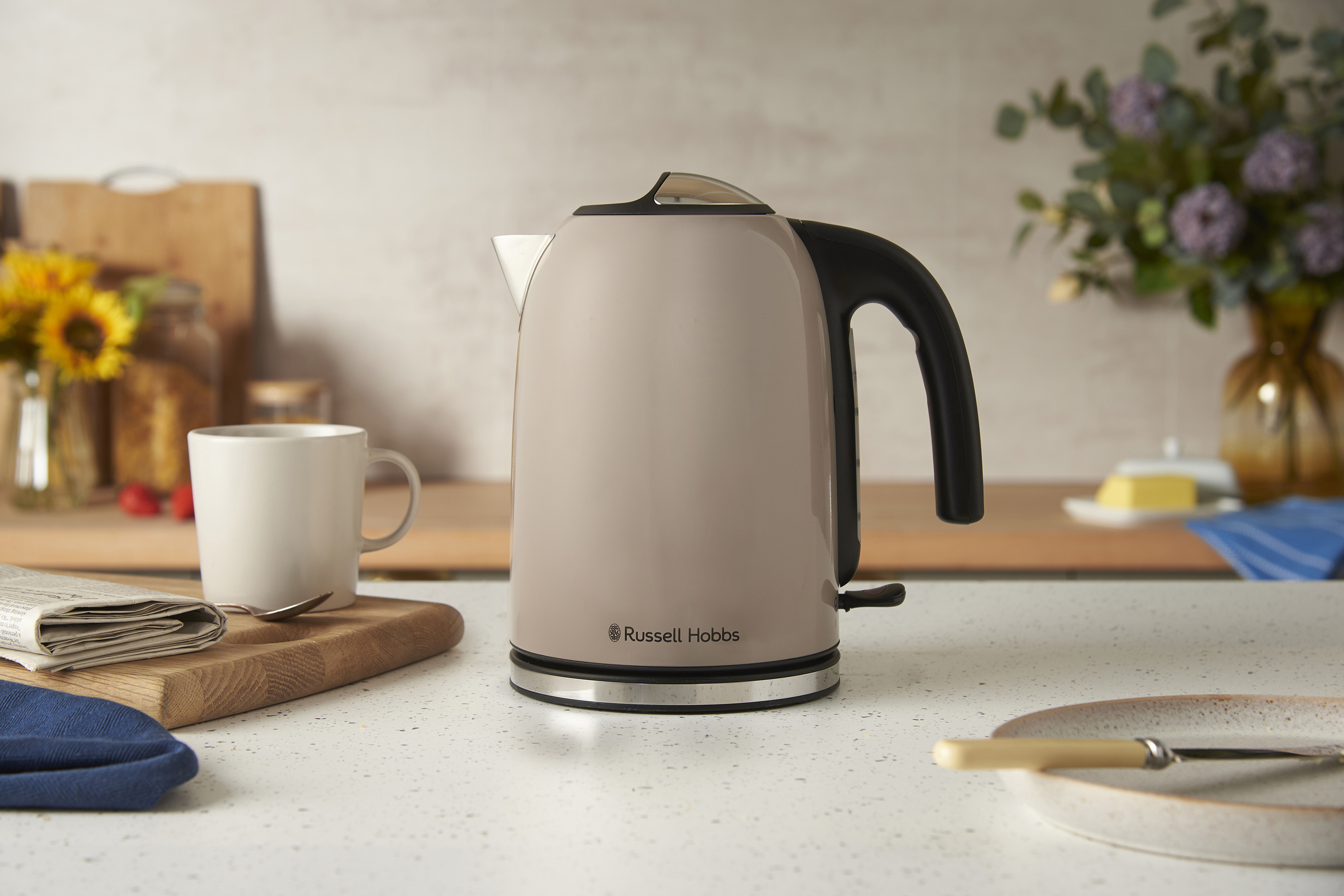 Egy bézs Russell Hobbs vízforraló a pulton. Egy bögre, újság és napraforgók a háttérben.