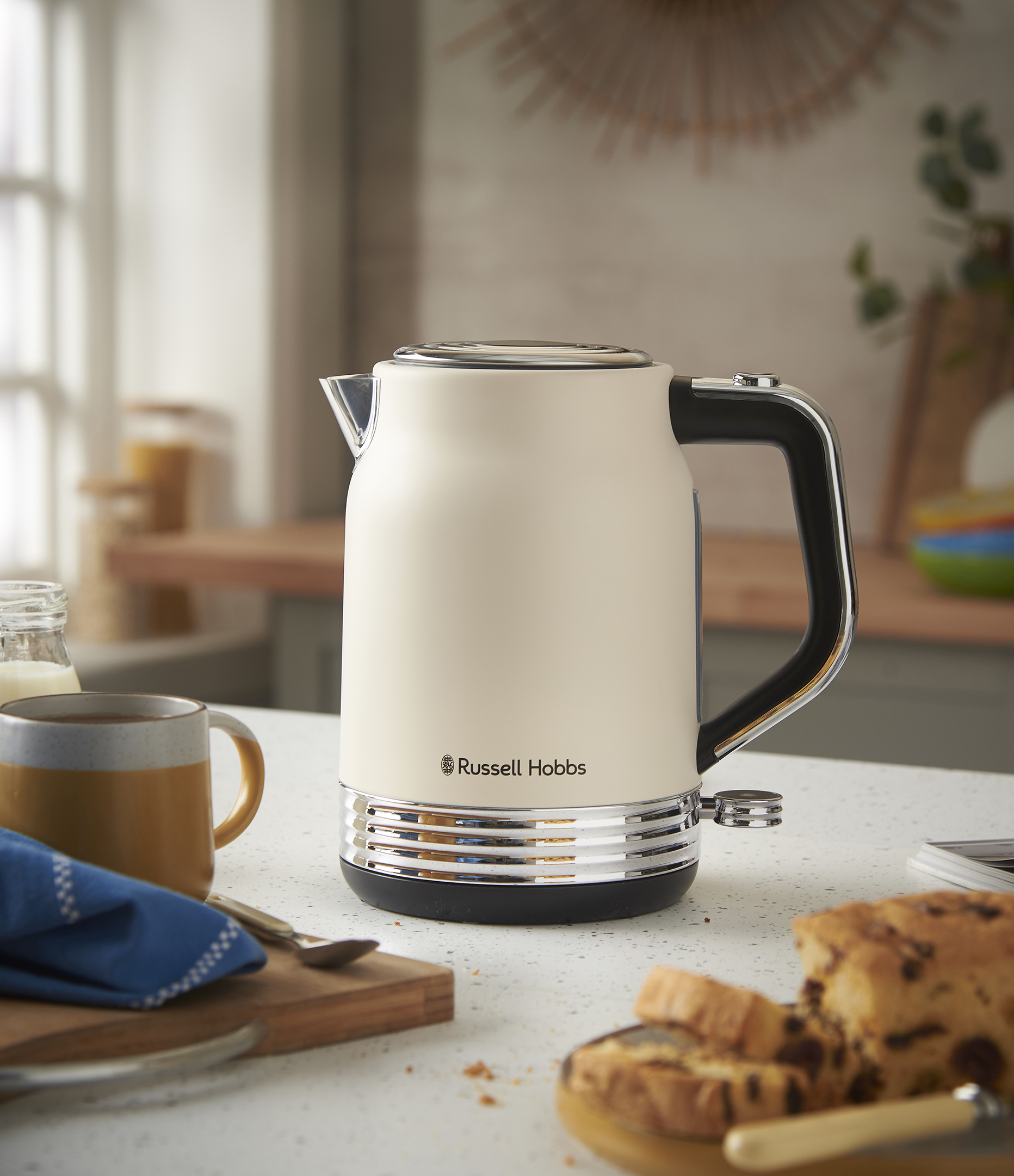 Krémszínű Russell Hobbs vízforraló pulton csészével, kenyérrel és tejjel. Konyhai környezet elmosódott háttérrel.