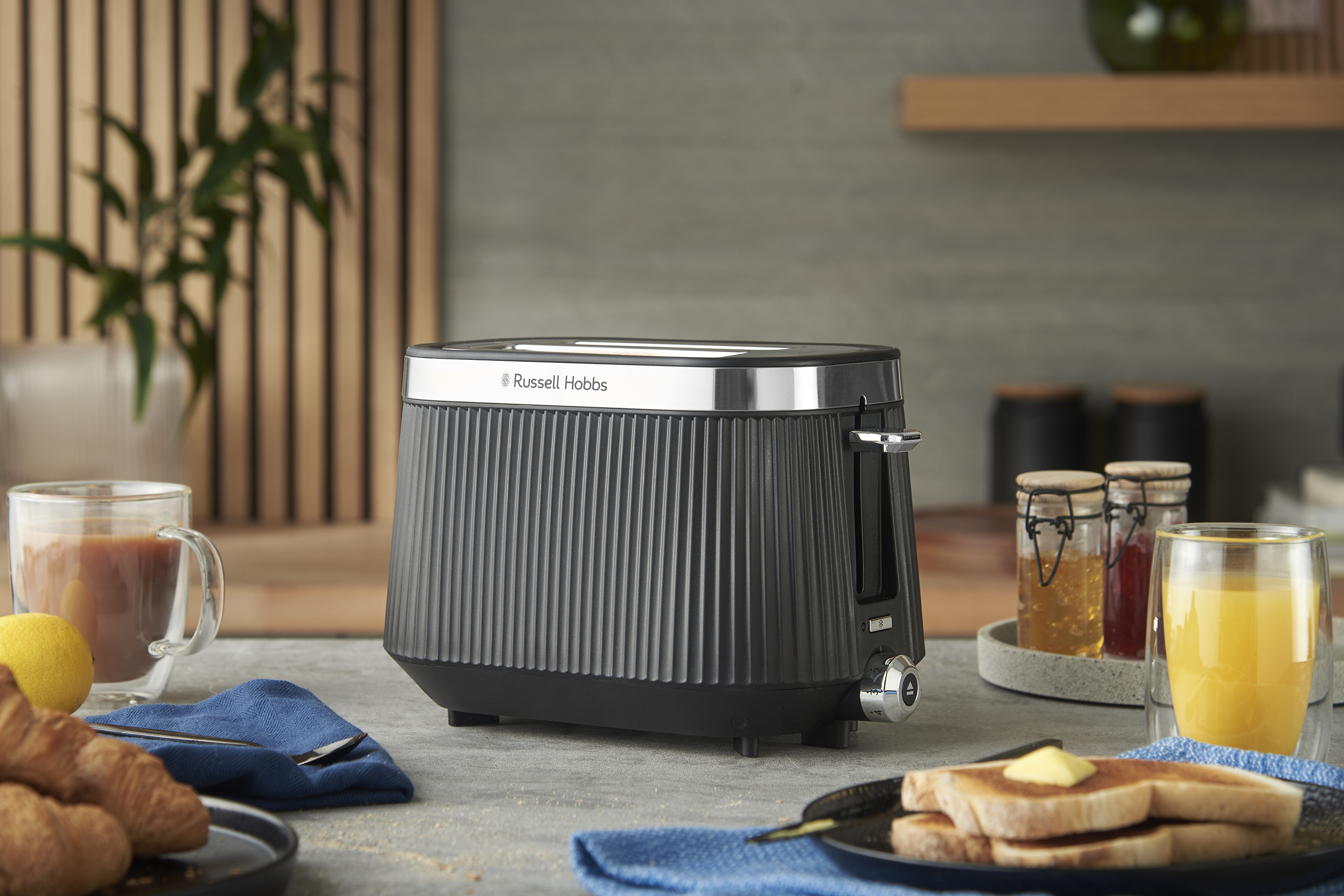 Egy fekete Russell Hobbs kenyérpirító pirítóssal, gyümölcslével és péksüteménnyel. A pirítóshoz lekváros üvegek, balra kávé.