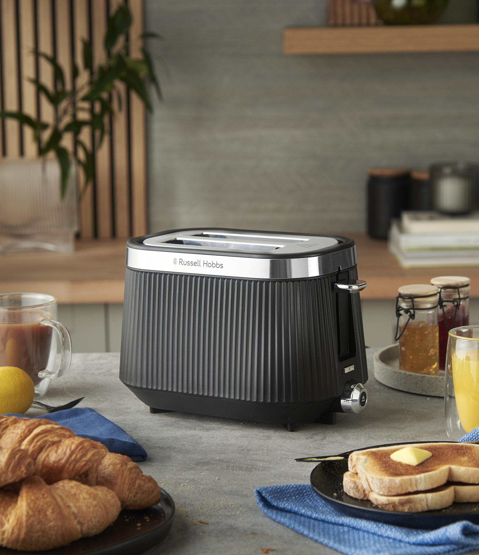 Egy fekete Russell Hobbs kenyérpirító egy asztalon áll. Pirítós, croissant és lekvár van a közelben. Egy csésze tea és narancslé is van.
