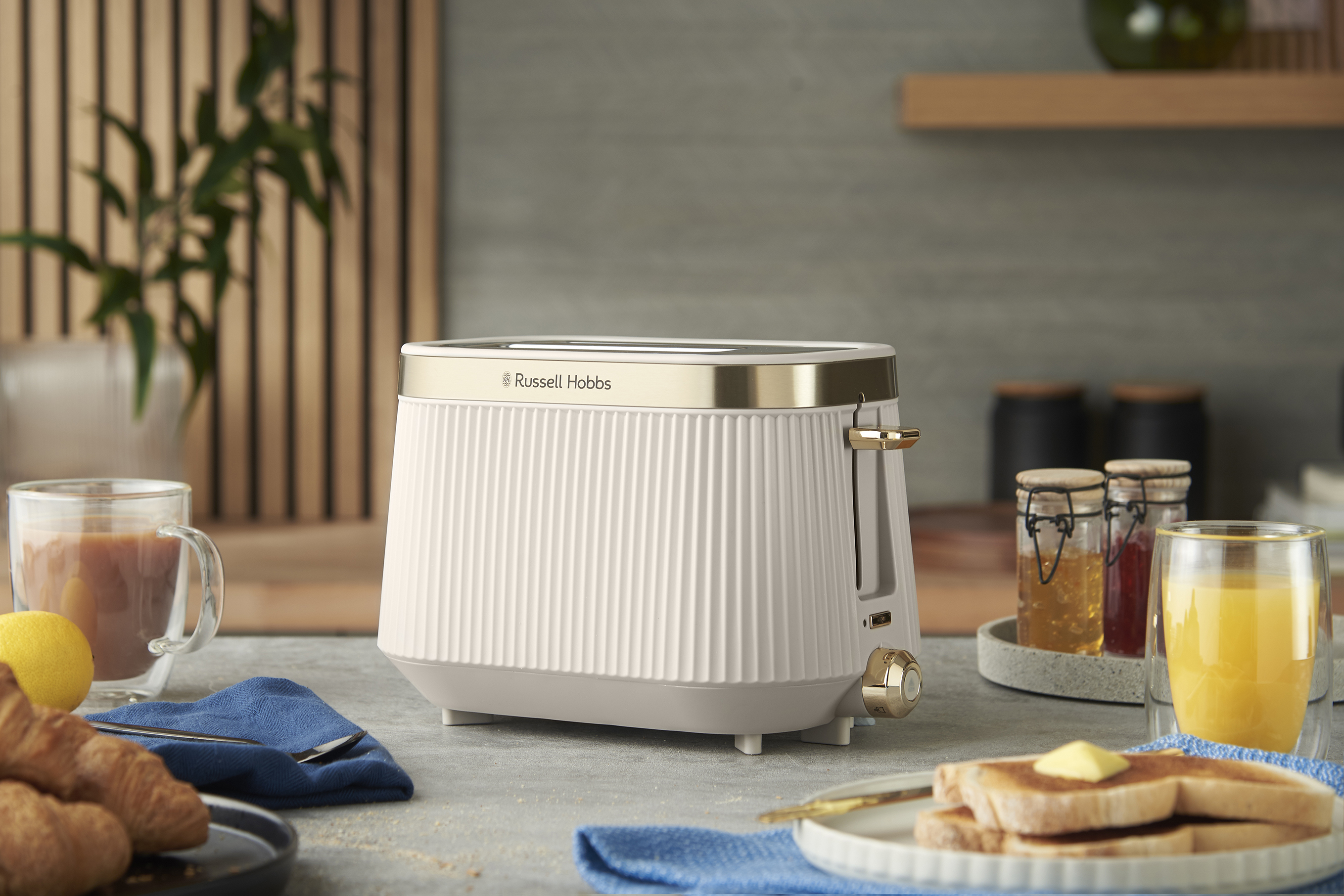 Egy fehér és arany Russell Hobbs kenyérpirító egy pulton van kenyérrel, lével és lekvárral. Mellette egy csésze kávé van.