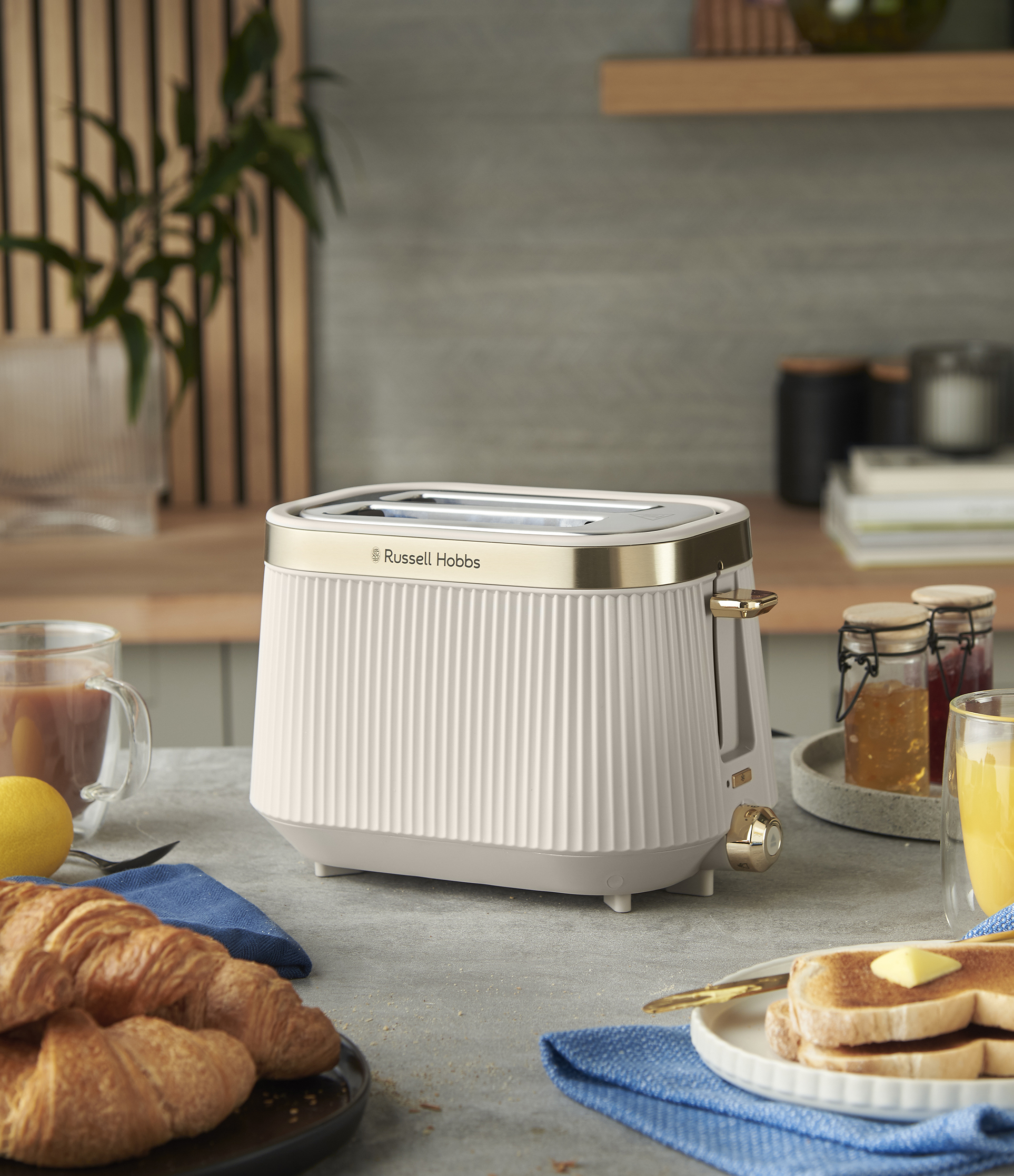 Egy krémszínű Russell Hobbs kenyérpirító, croissant, vajjal pirított kenyér és gyümölcslé van egy asztalon. Láthatók lekváros üvegek és kávé.