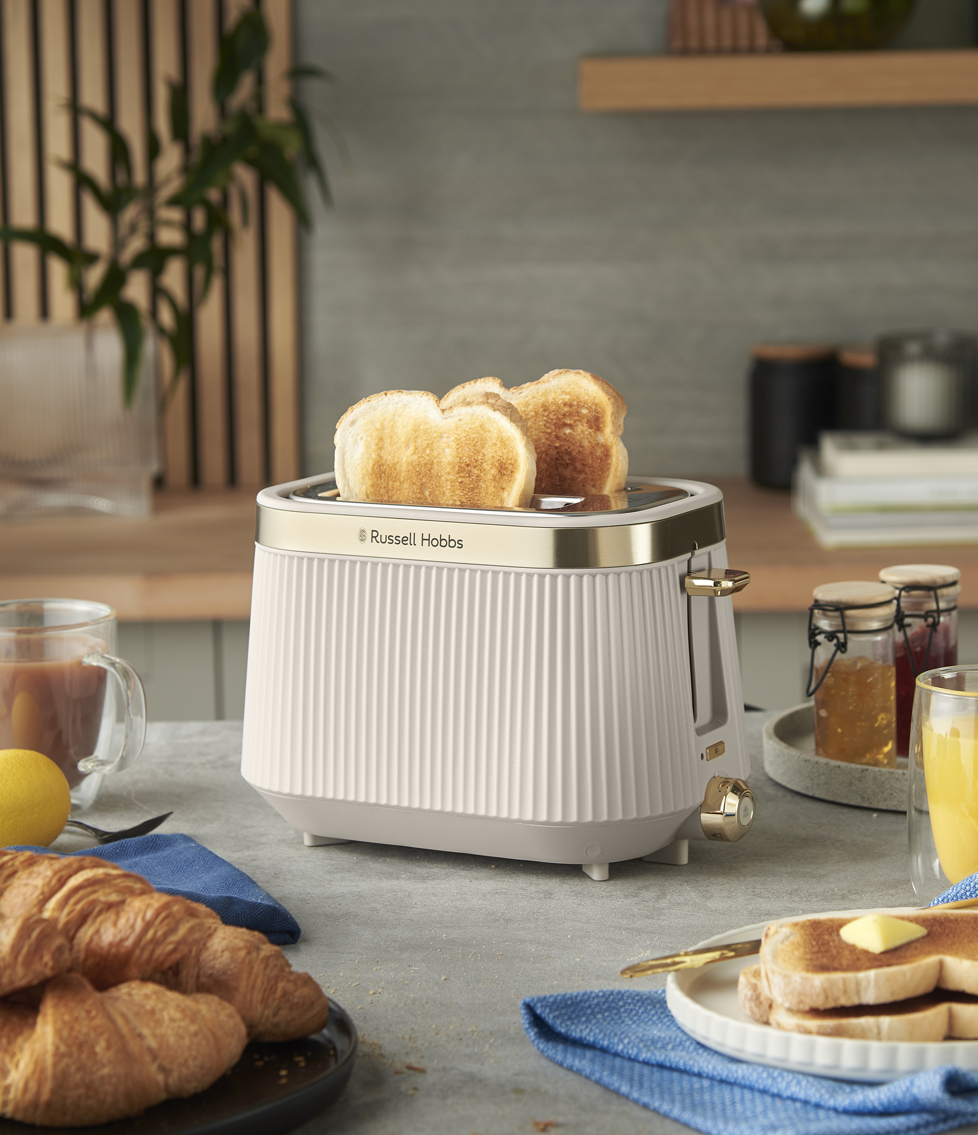 Egy kenyérpirító pirítóssal, croissant-okkal és gyümölcslével a pulton. Dzsemes üvegek, citrom és egy csésze kávé is látható.