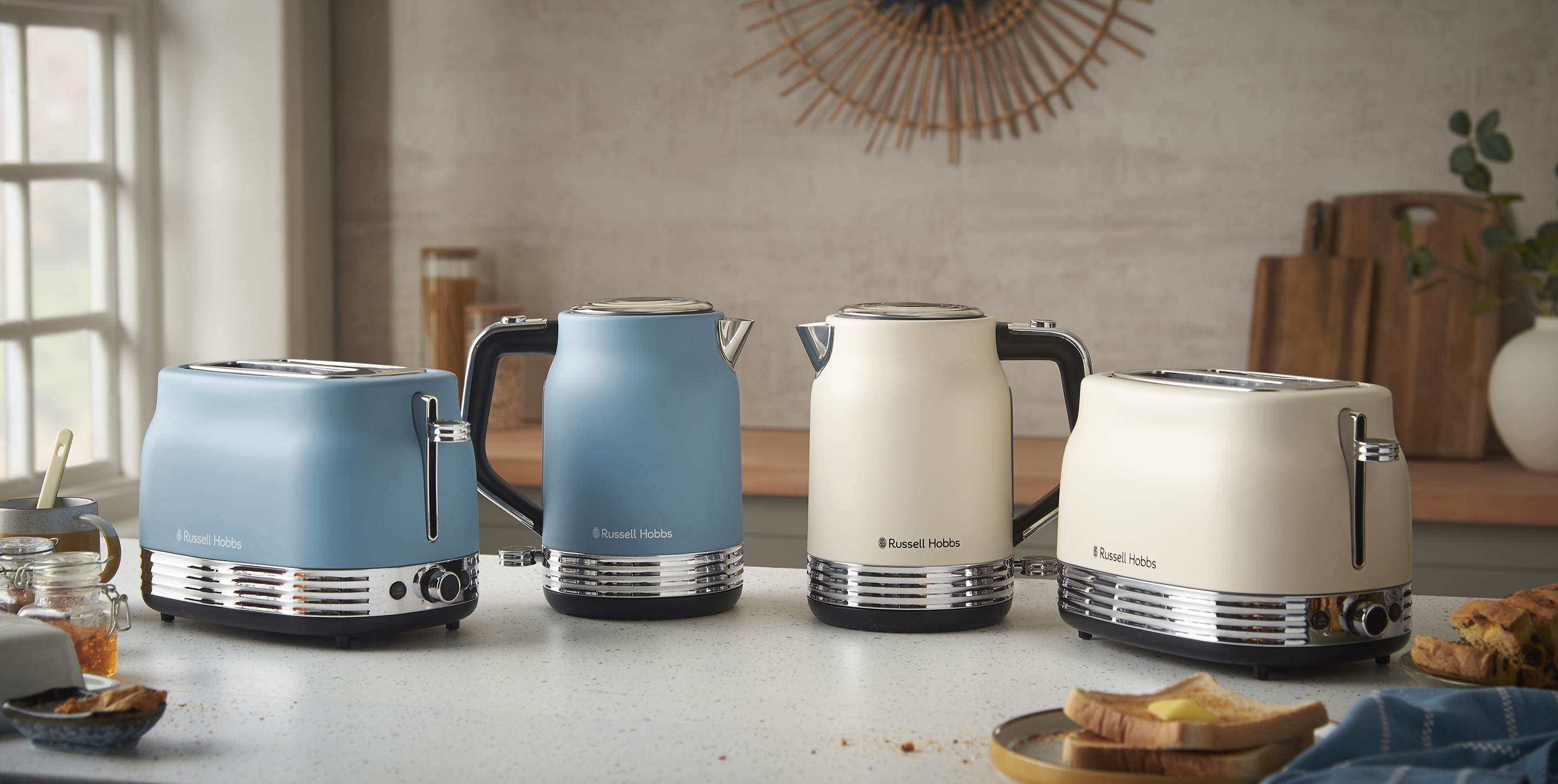 Négy Russell Hobbs konyhai eszköz: két kenyérpirító és két vízforraló, kék és krémszínben, fehér pulton, ablak közelében.