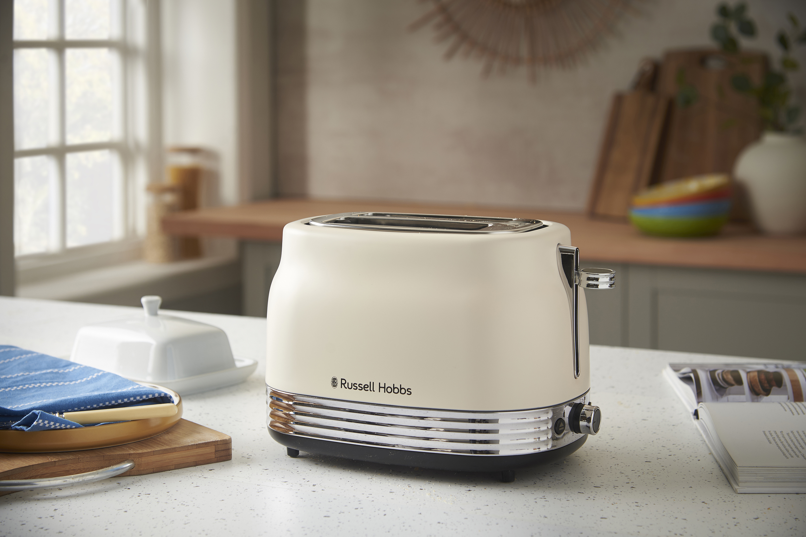 Krémszínű Russell Hobbs kenyérpirító a pulton. Vajtartó, ablak és vágódeszkák a háttérben.