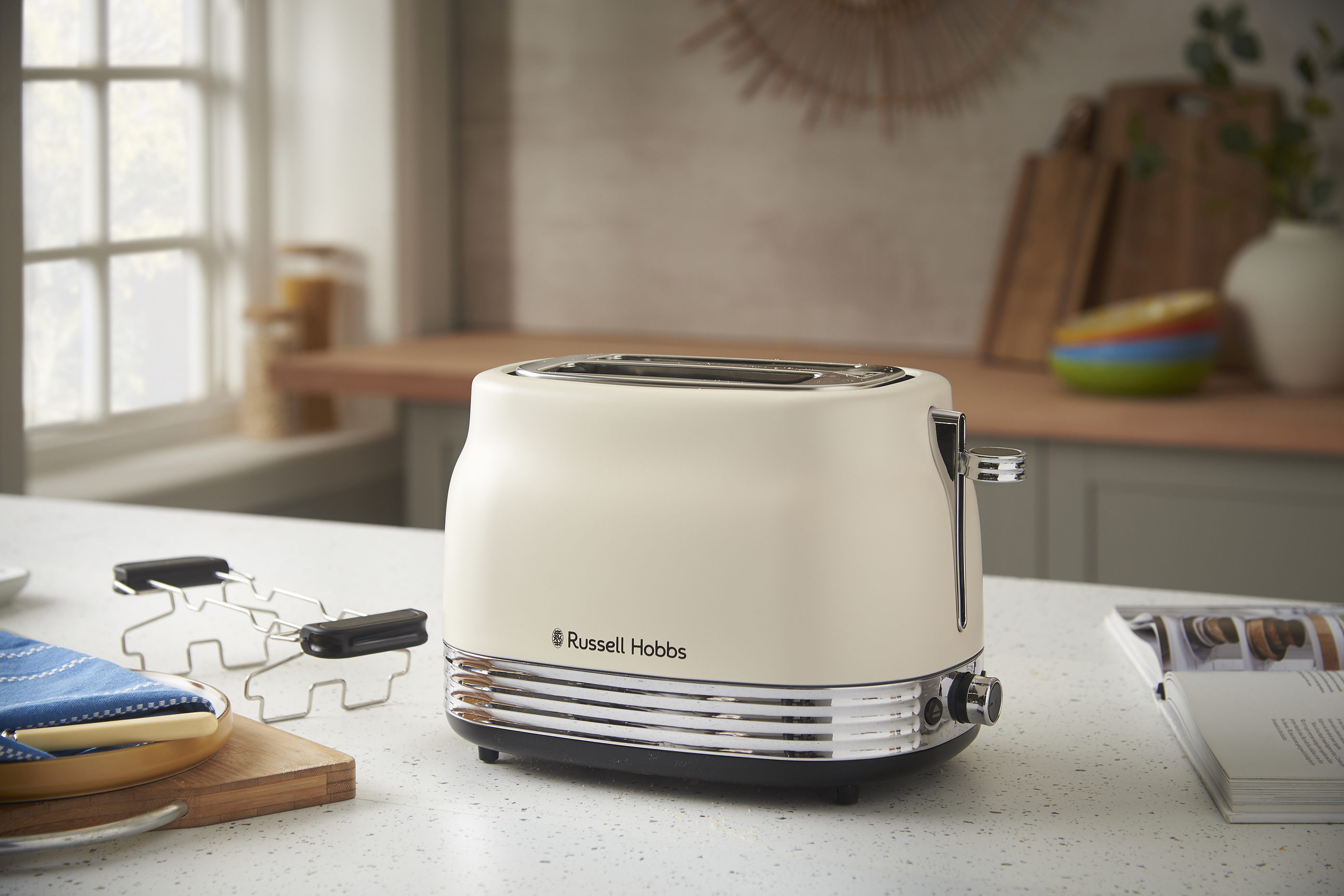 Egy krémszínű Russell Hobbs kenyérpirító a pulton. Egy tányér, egy vágódeszka és egy ablak a jelenetben.