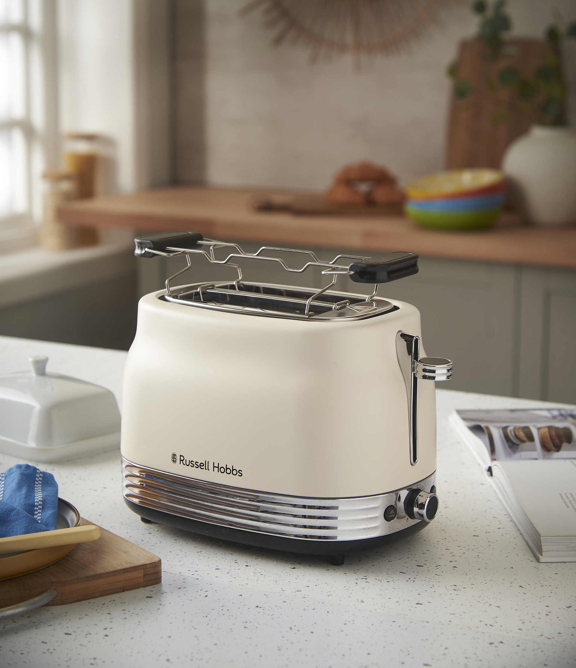 Krémszínű Russell Hobbs kenyérpirító a pulton. Vajtartó, fa deszka és tálak közelben. Konyhai háttér.