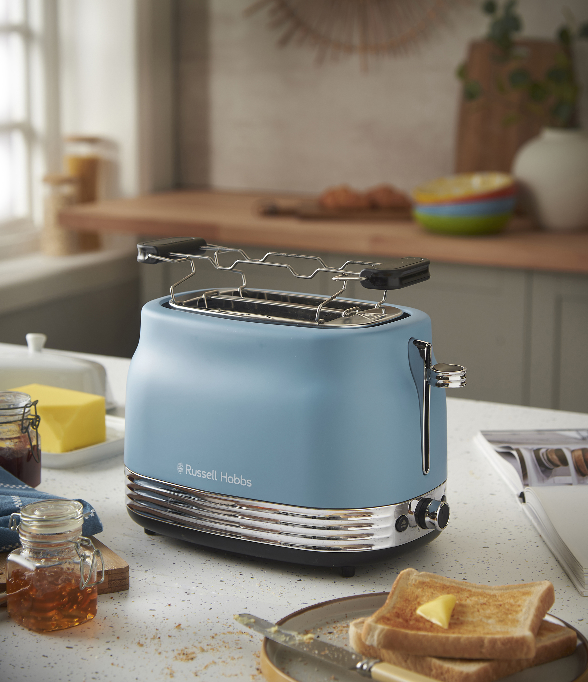 Egy kék Russell Hobbs kenyérpirító egy pulton áll, pirítóssal, vajjal, lekvárral és mézzel. A háttérben egy ablak és konyhai elemek láthatók.