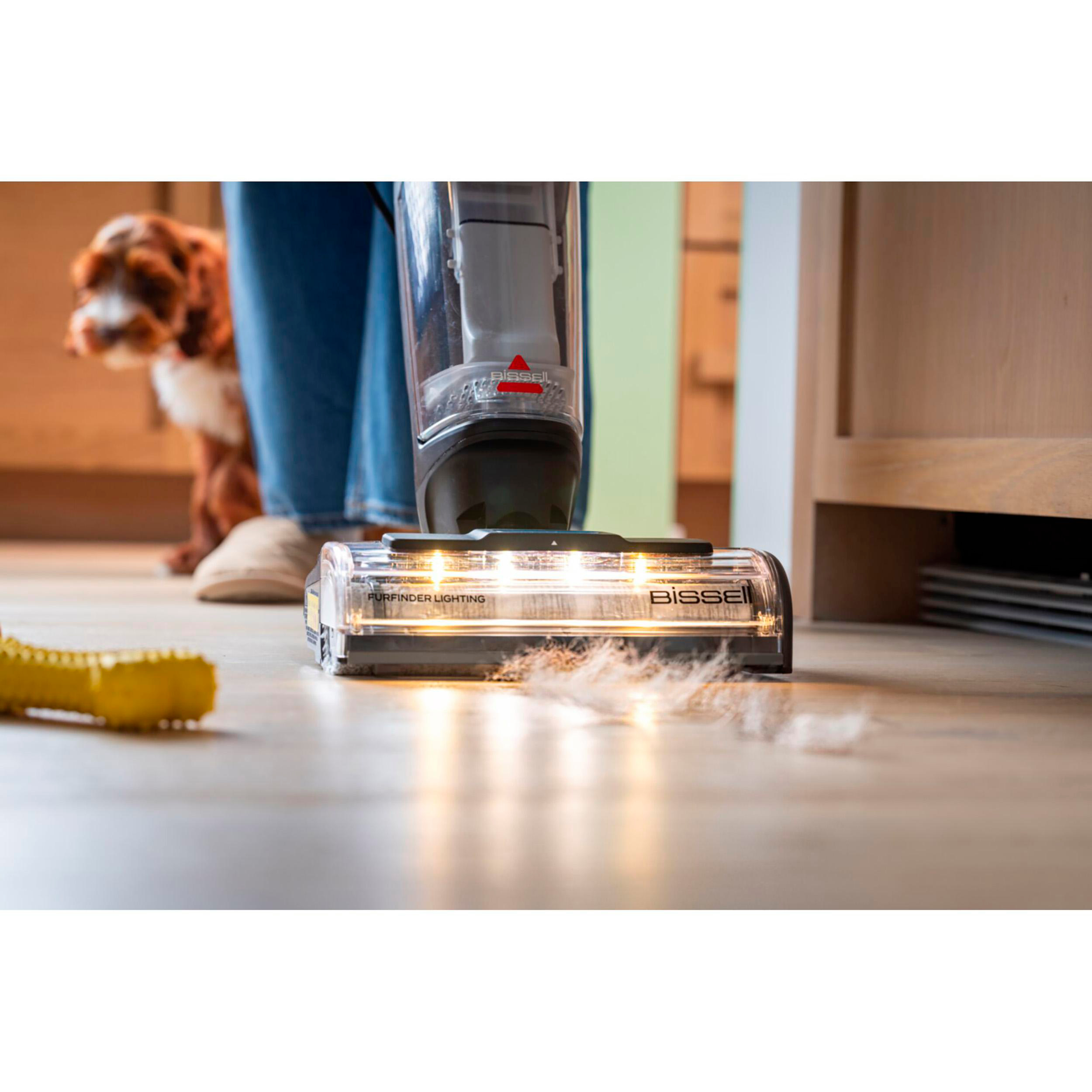 Eine Person saugt den Boden mit einem Bissell-Staubsauger, im Hintergrund ist ein Hund zu sehen, und ein gelbes Spielzeug liegt auf dem Boden.