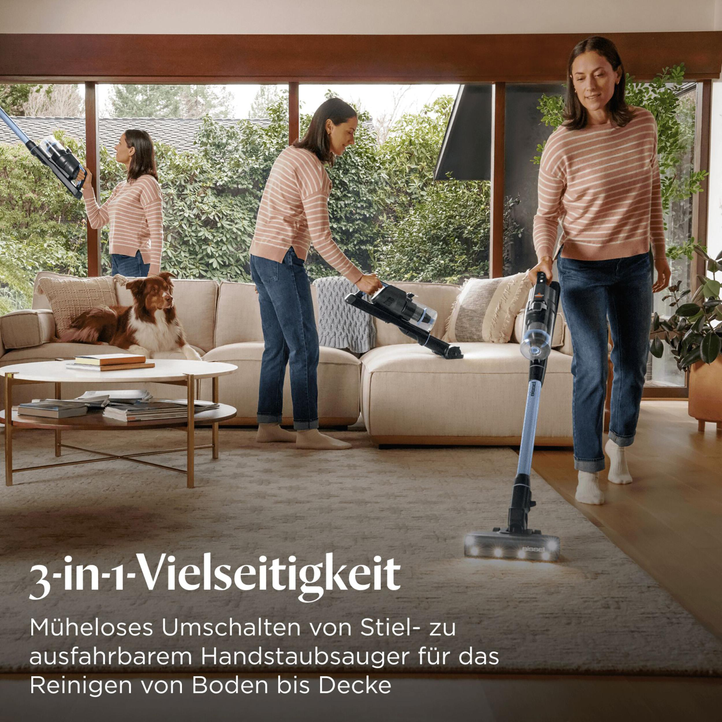 Eine Frau saugt einen Teppich mit einem kabellosen Staubsauger. Eine andere Frau saugt das Sofa. Ein Hund liegt auf der Couch.