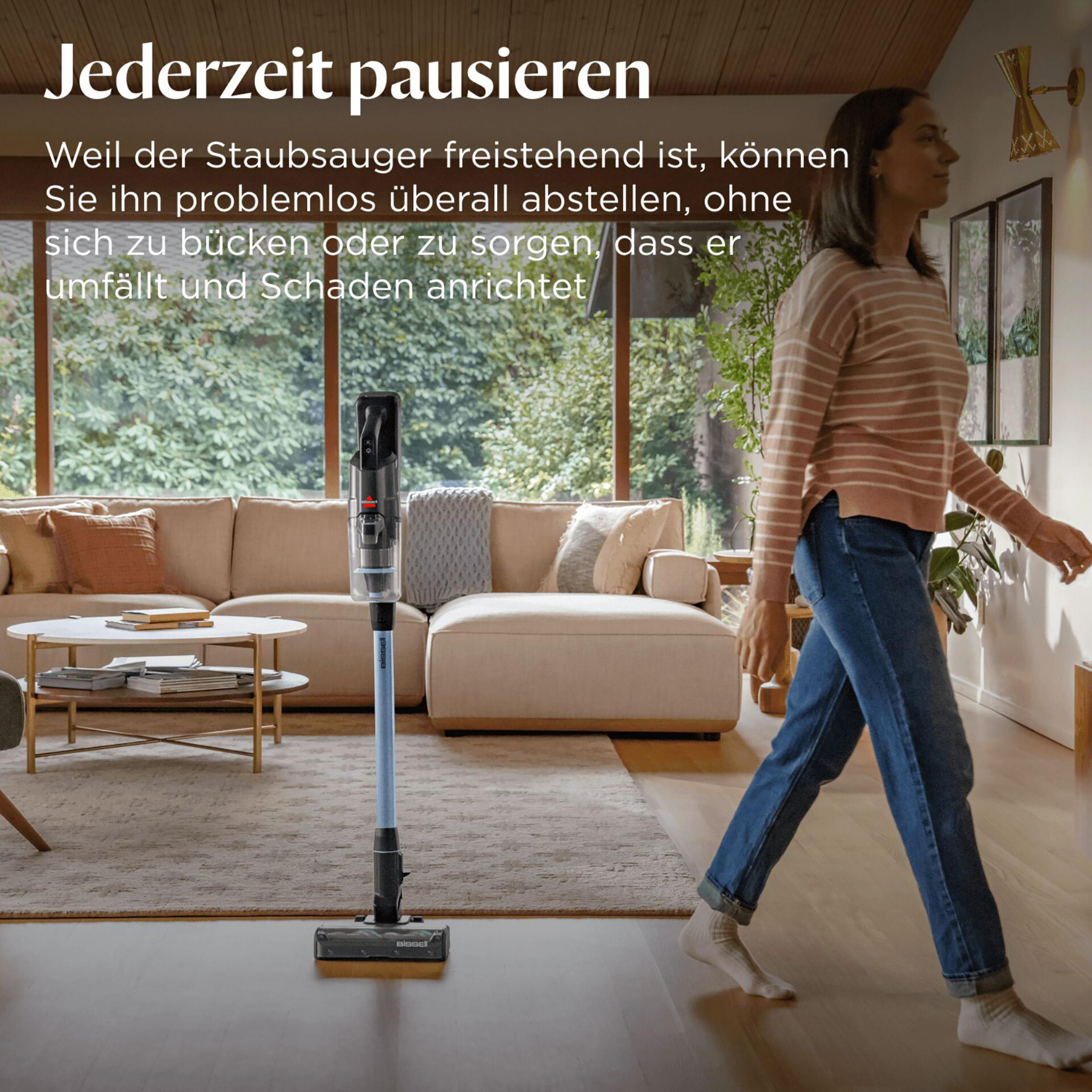 Eine Frau geht an einem Staubsauger vorbei. Der Staubsauger steht aufrecht. Die Frau trägt Jeans und ein gestreiftes Hemd.