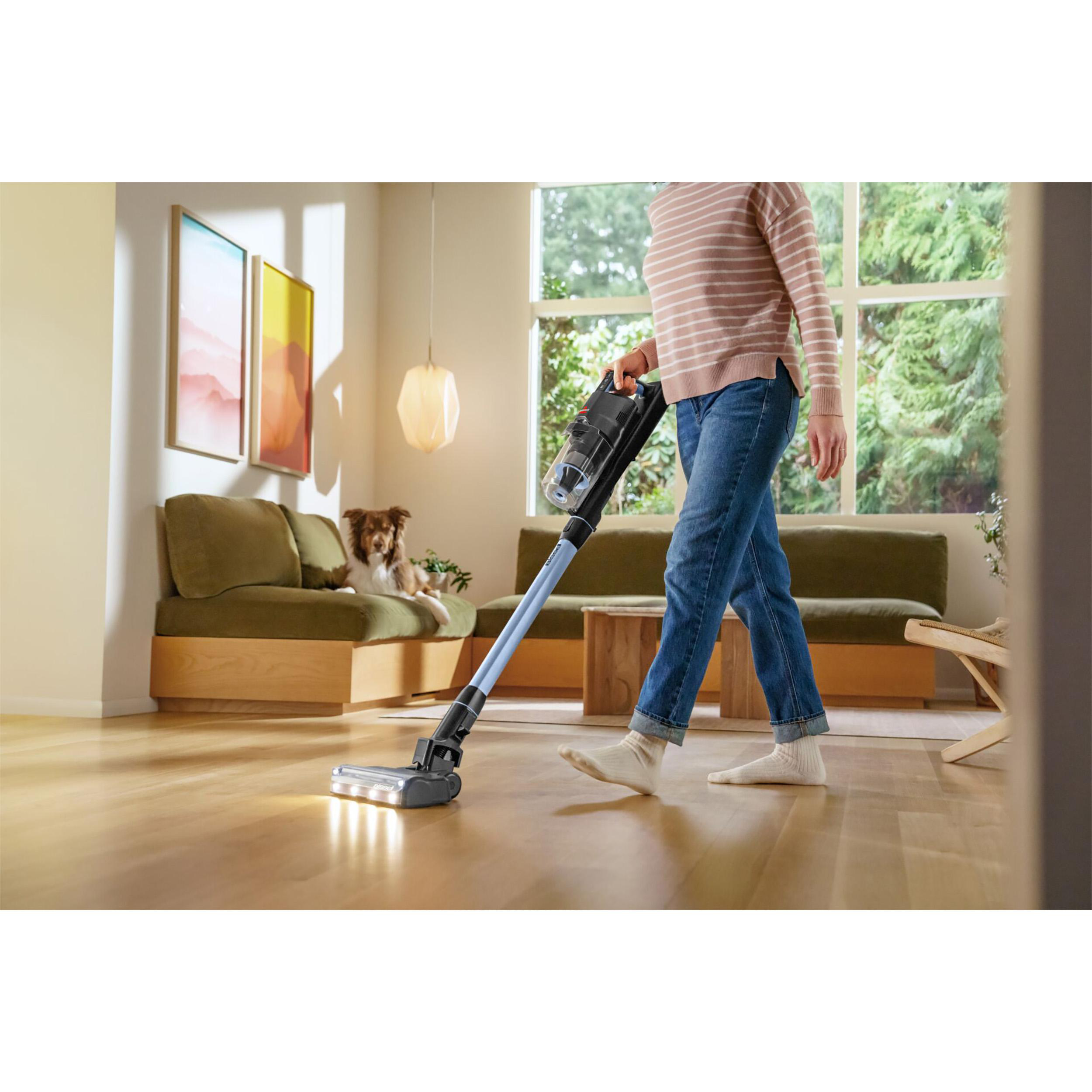 Eine Person saugt einen Raum mit einem kabellosen Staubsauger. Ein Hund ist im Hintergrund auf einem Sofa.