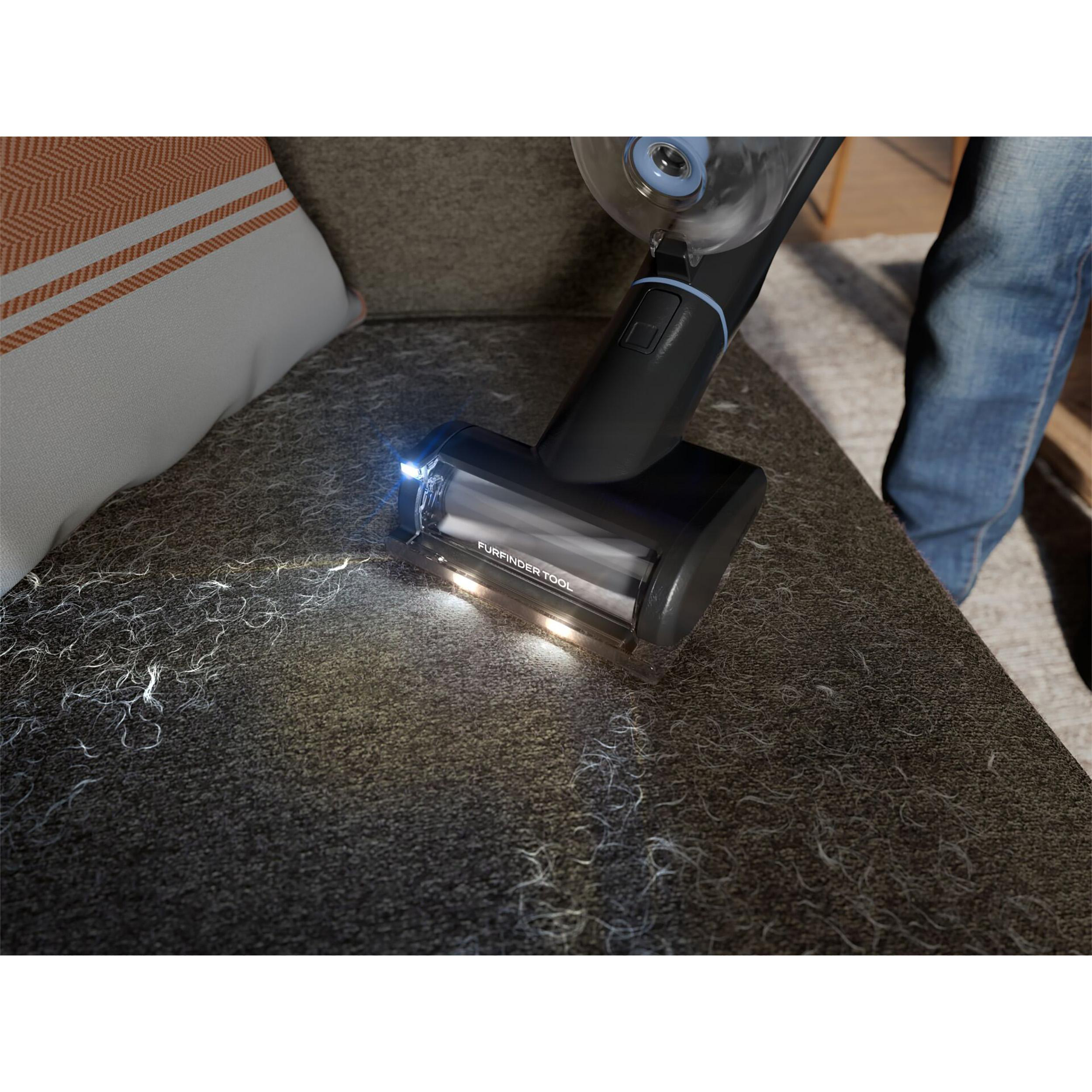 Ein Staubsauger mit Licht reinigt ein graues Sofa mit Tierhaaren und einem gemusterten Kissen.