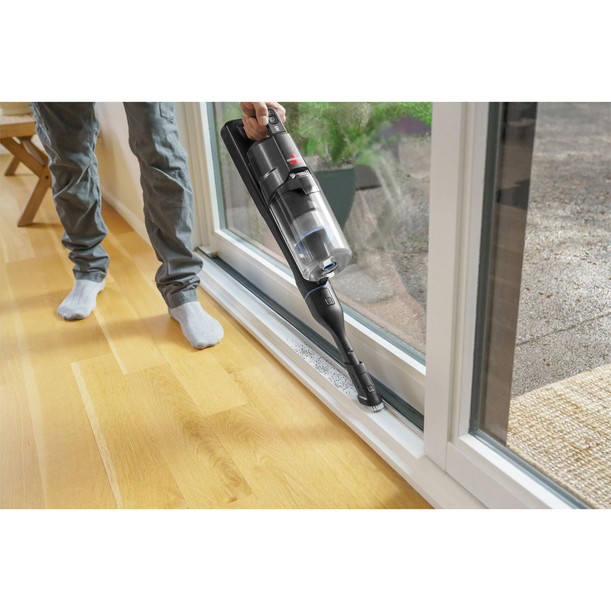 Person saugt einen Fensterrahmen. Ein schwarzer Staubsauger wird an einem Fenster verwendet. Holzboden und Außenbereich sind sichtbar.