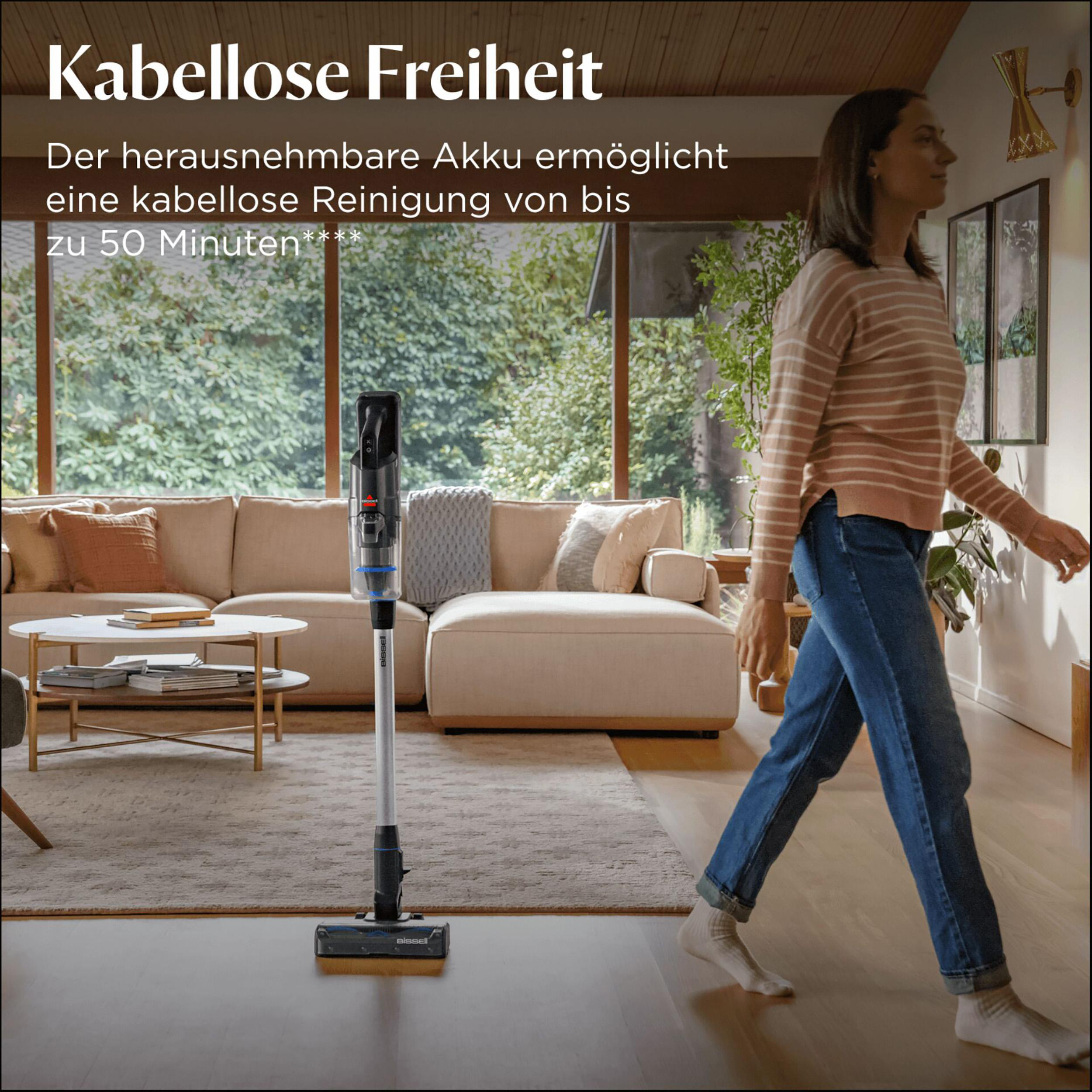 Eine Frau in einem gestreiften Hemd geht an einem Staubsauger vorbei. Wohnzimmer mit Sofa und Fenster im Hintergrund.