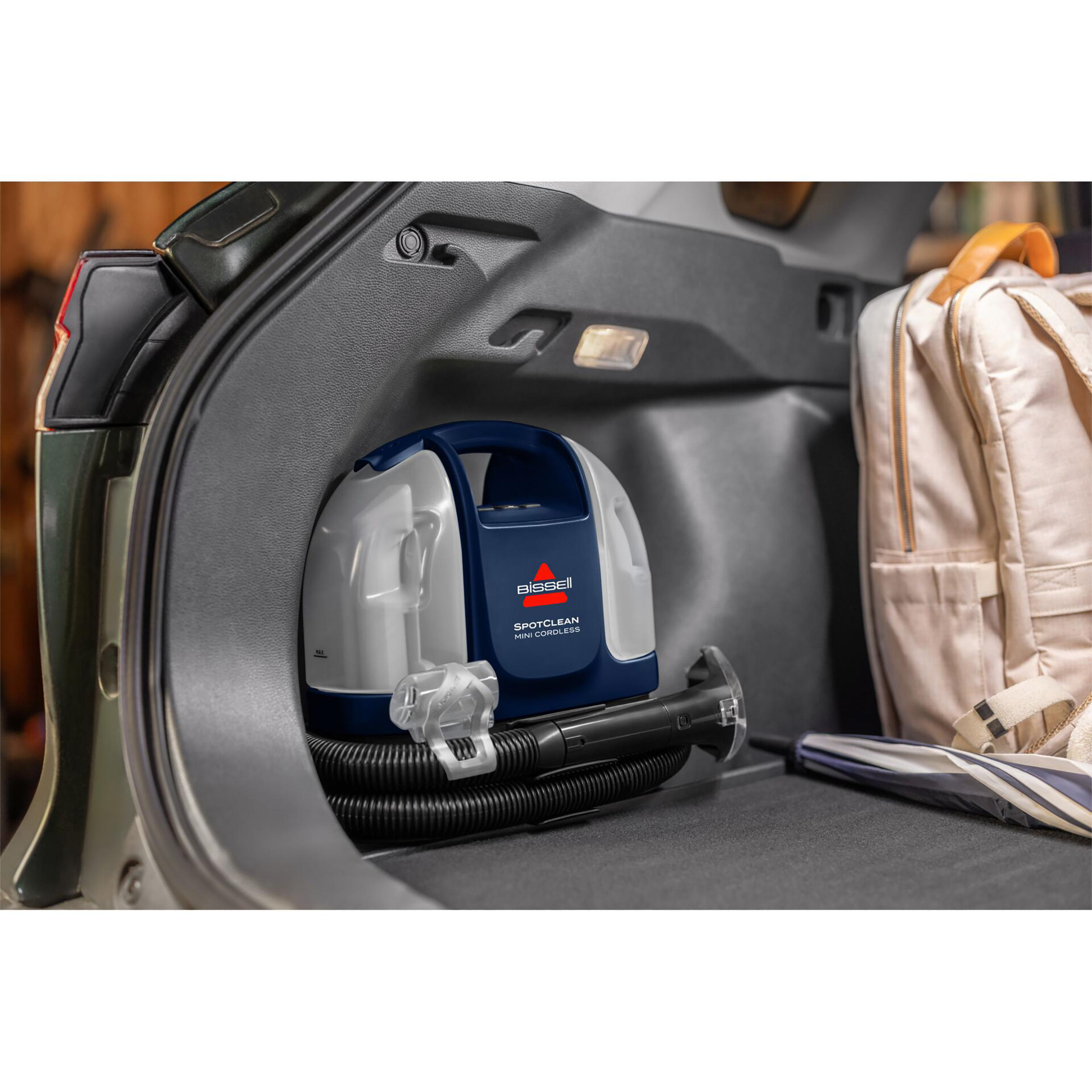 Ein tragbarer Bissell SpotClean Mini Cordless-Reiniger befindet sich in einem Kofferraum. Rucksack und Schirm sind daneben.