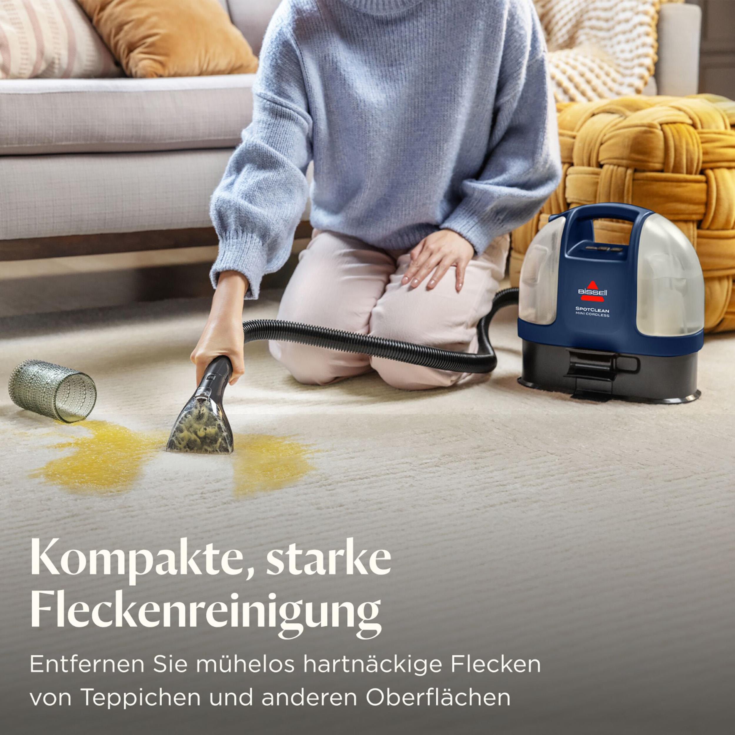 Eine Frau reinigt einen Teppichfleck mit einem Bissell SpotClean. Im Hintergrund sind ein Sofa und ein gewebter Hocker.
