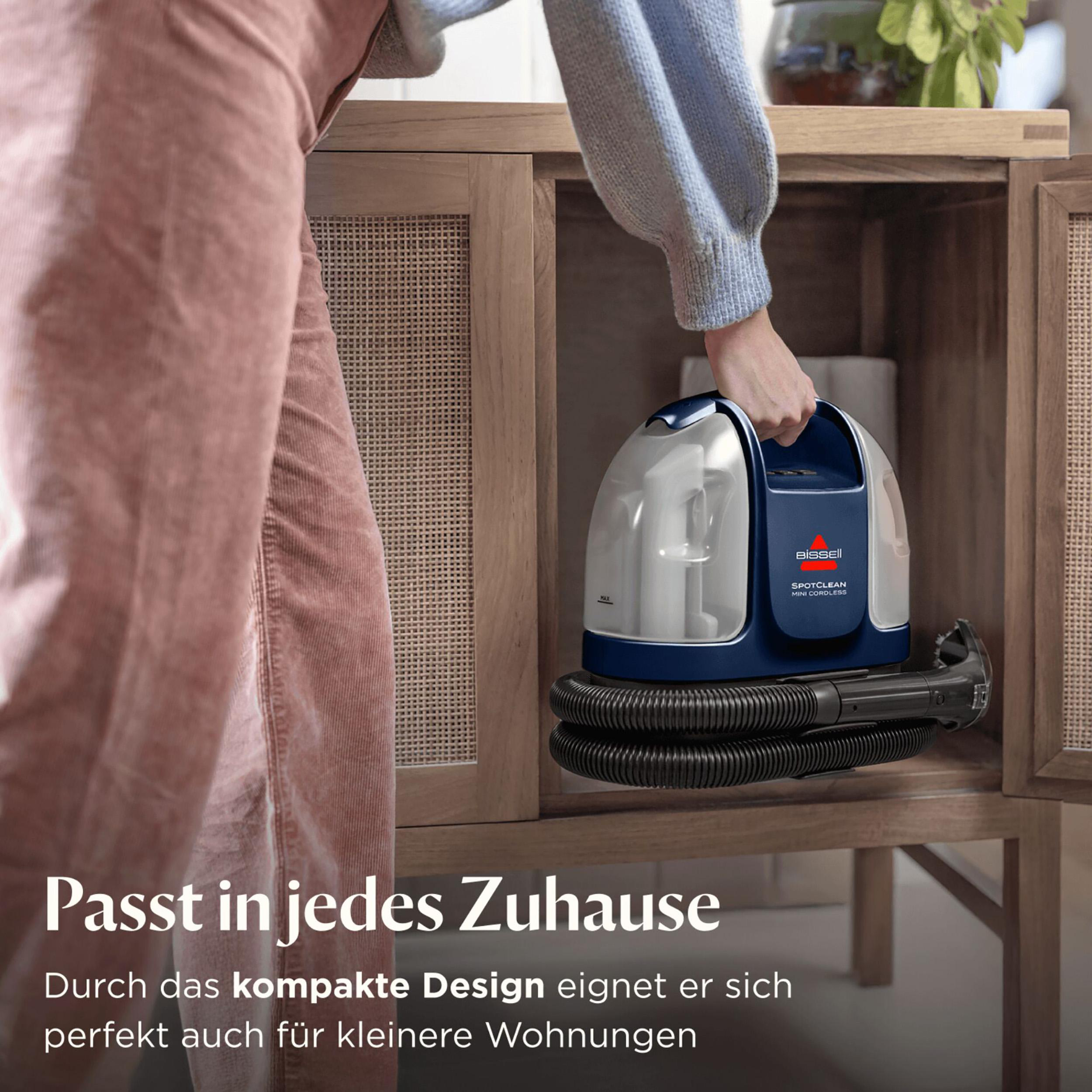 Eine Person hält einen blau-weißen Bissell SpotClean Mini Cordless Staubsauger in einem Holzschrank.