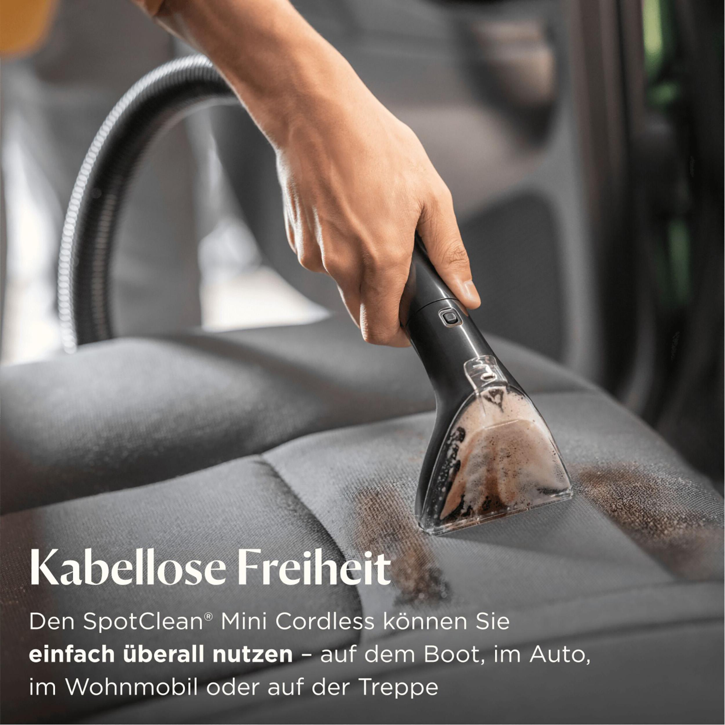Eine Hand benutzt einen schwarzen Staubsaugerkopf auf einem grauen Autositz, um einen Fleck zu entfernen. Der Text lautet 'Kabellose Freiheit'.