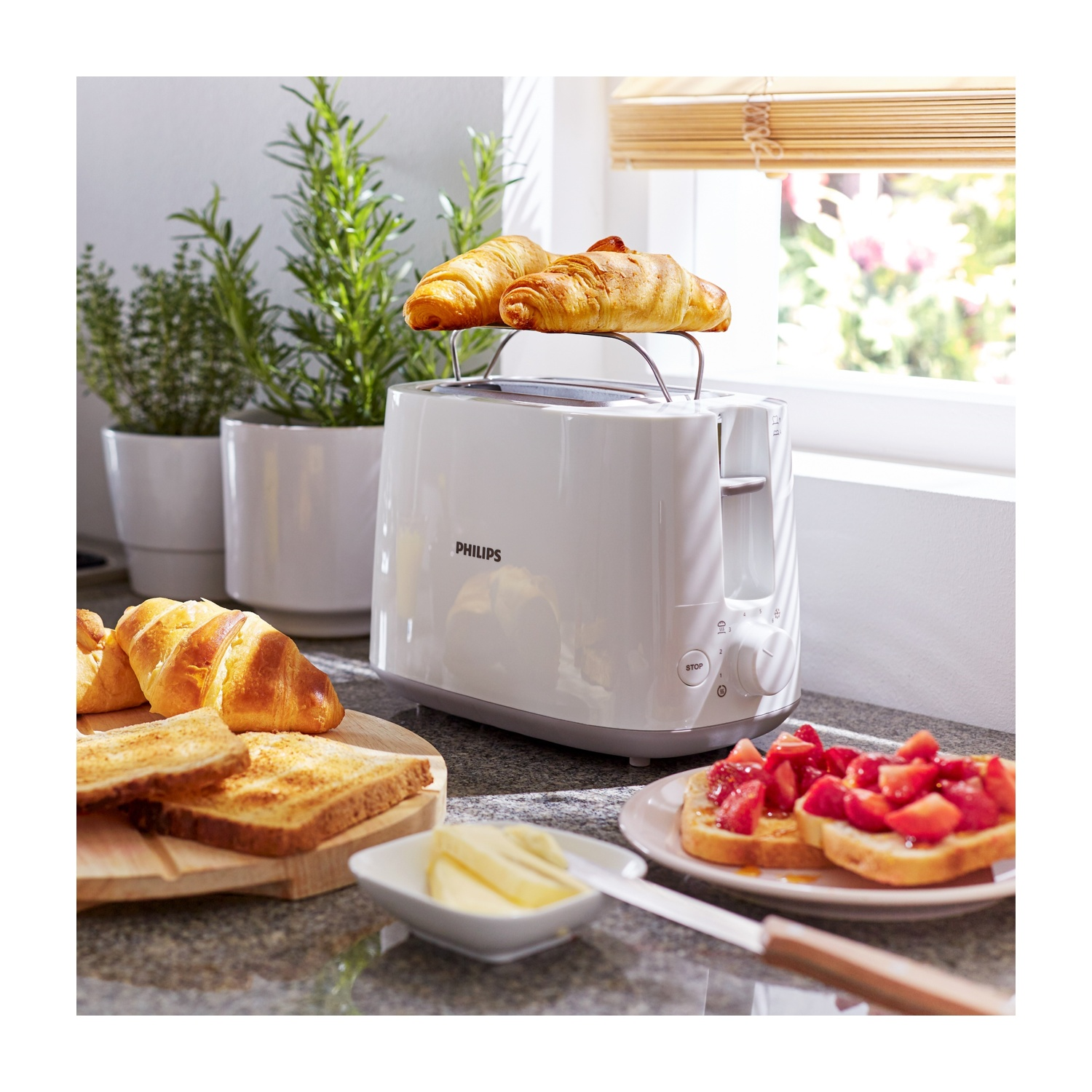 Un tostapane Philips bianco con croissant sopra. Toast, burro e toast alle fragole sono sul bancone, con piante sullo sfondo.