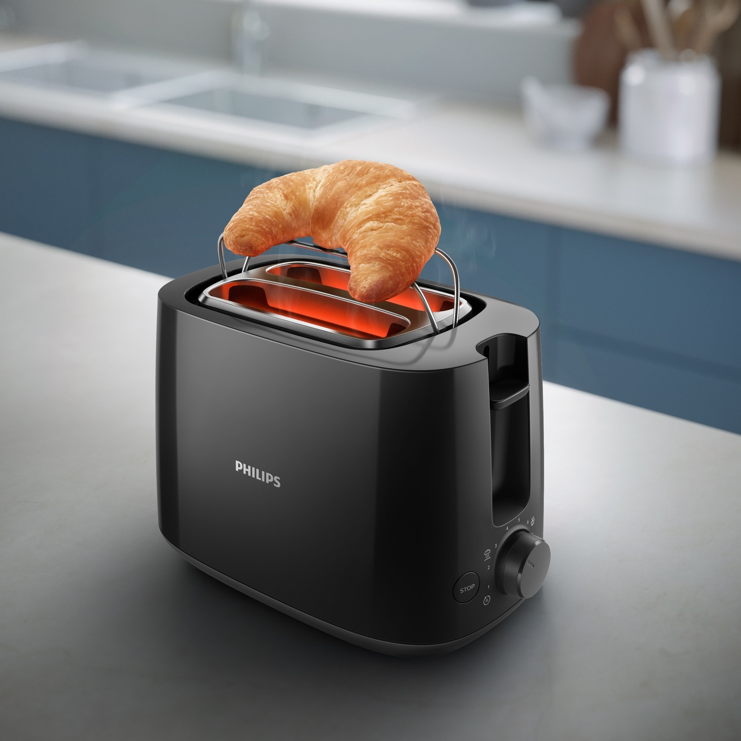 Un tostapane Philips nero con un croissant dorato sopra. Il tostapane è su una superficie grigia, con una cucina blu e bianca sullo sfondo.
