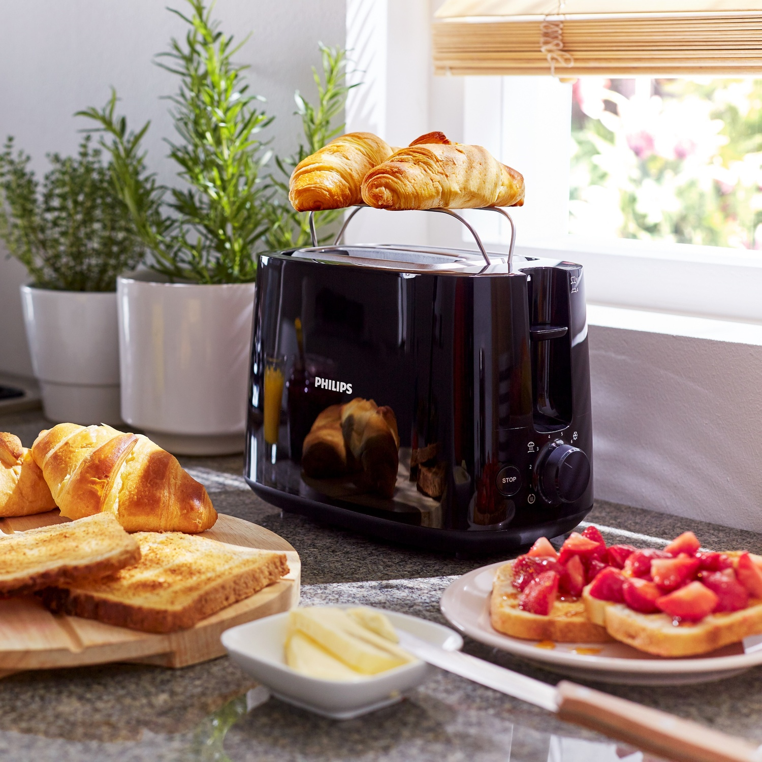 Un tostapane Philips nero con croissant sopra. Toast, fragole, burro e un coltello sul bancone.