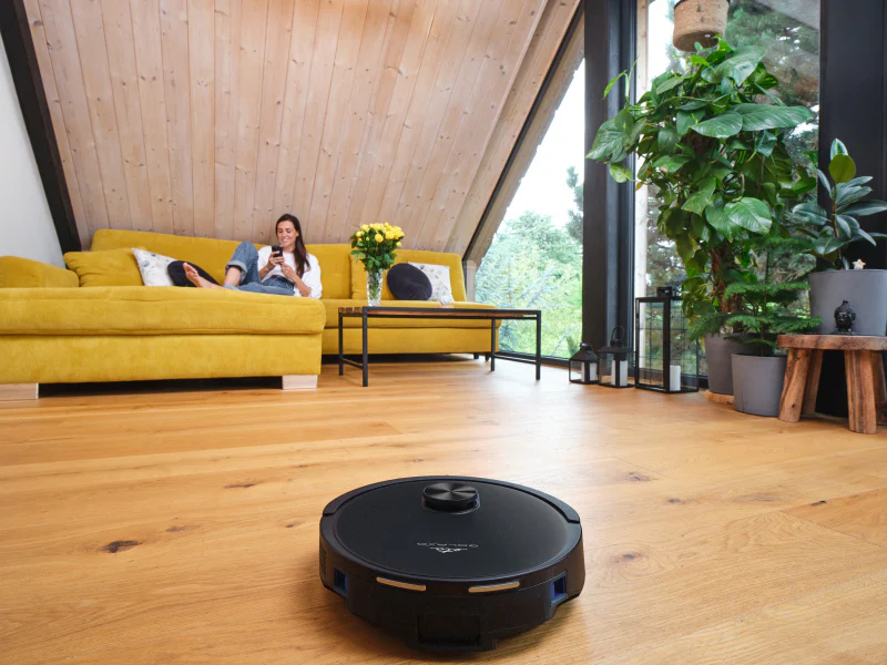 Egy nő egy sárga kanapén a telefonját használja, miközben egy robotporszívó tisztítja a fa padlót. Növények és egy ablak láthatók.