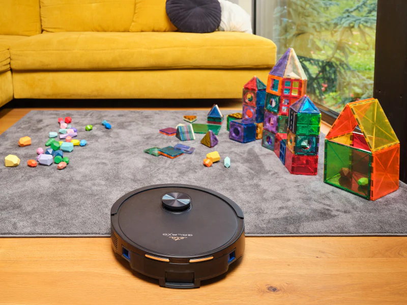 Egy robot porszívó egy szürke szőnyegen. Színes játékkockák szanaszét. Egy sárga kanapé a háttérben.