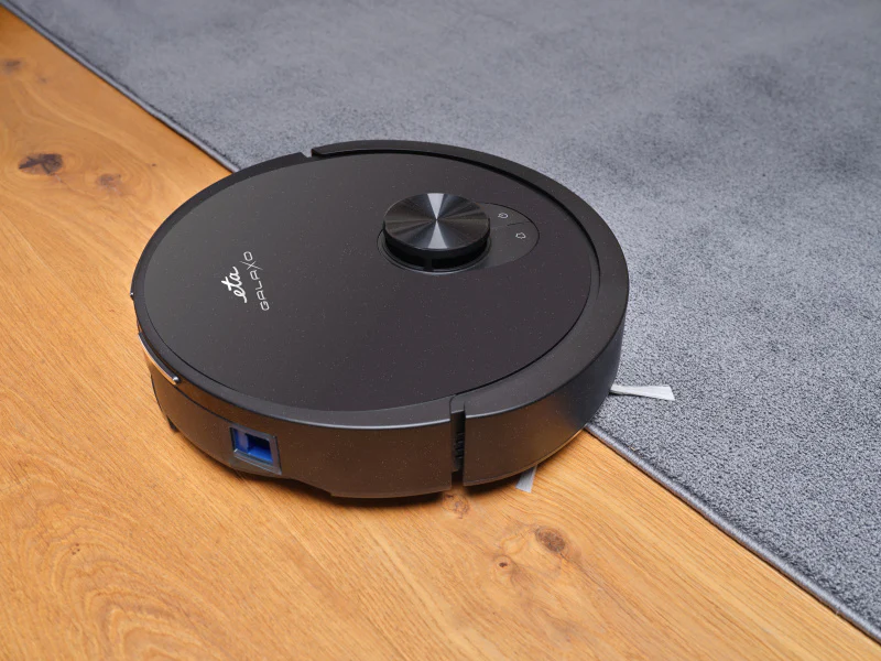 Egy fekete robotporszívó a fa padlón és a szürke szőnyegen. A porszívó a szőnyeg szélénél van, a márkanév látható.