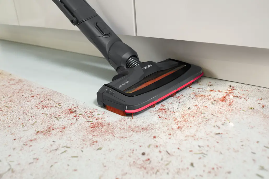 Egy szürke és piros Philips porszívó tisztít egy fehér padlót, melyen piros és zöld részecskék szóródnak, egy fehér szekrény alatt.