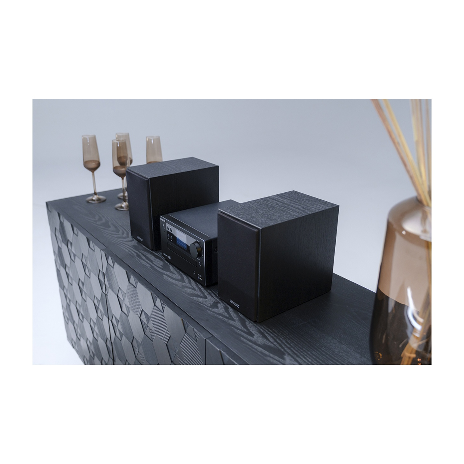Un impianto stereo nero con altoparlanti, calici da champagne e un vaso marrone con canne su un mobile in legno scuro.