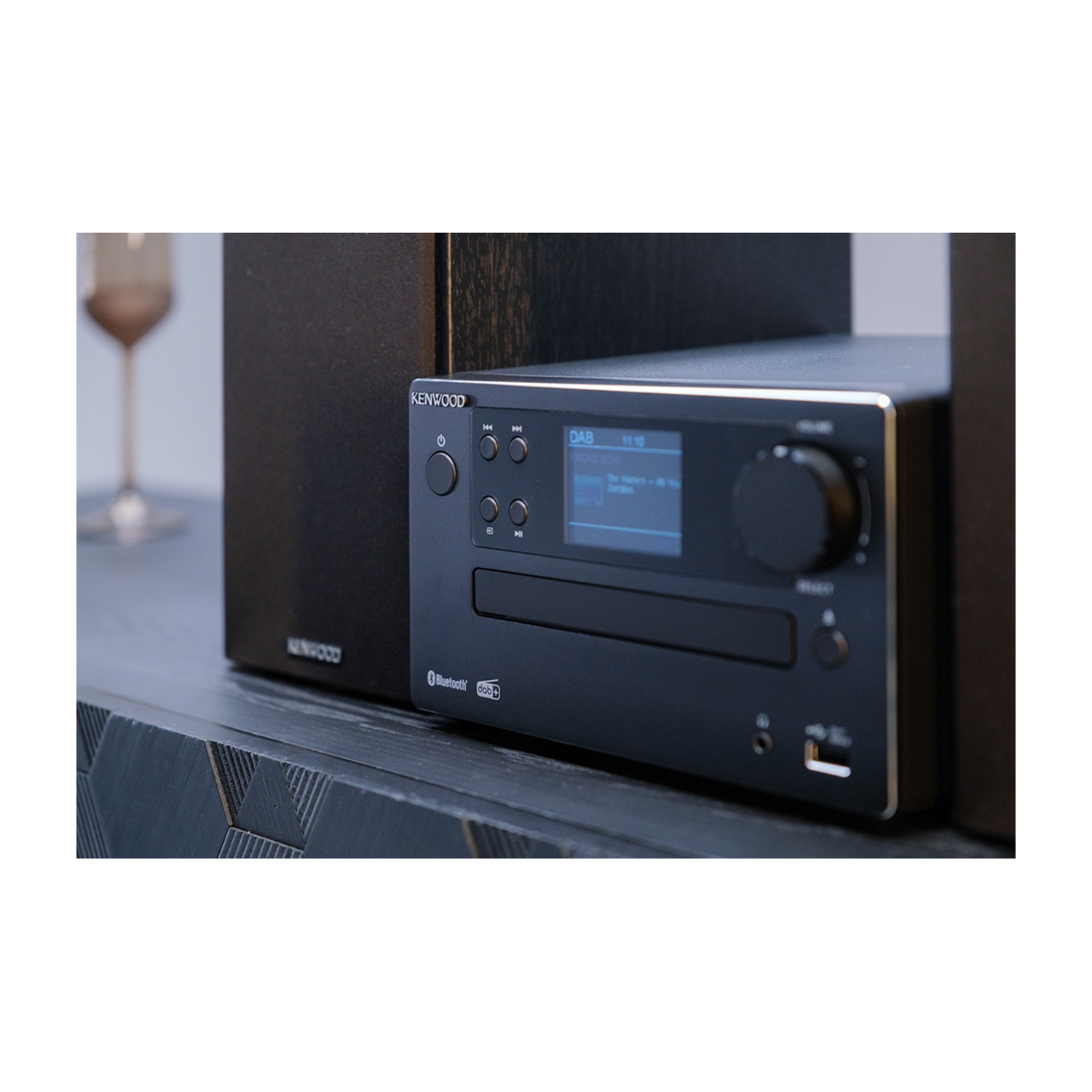 Un impianto stereo Kenwood nero con altoparlanti. Un bicchiere di vino sullo sfondo, lo stereo è su una superficie di legno scuro.