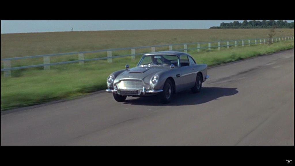 Silberner Aston Martin DB5 auf einer Straße. Grünes Feld, weißer Zaun und Bäume im Hintergrund. Klarer blauer Himmel.