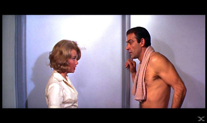 Eine Frau und ein Mann ohne Hemd mit einem Handtuch unterhalten sich in der Nähe einer Dusche. Die Frau trägt ein weißes Hemd.