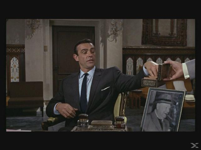 Sean Connery im Anzug nimmt eine Schachtel von einer anderen Person entgegen. Auf dem Tisch steht ein gerahmtes Foto eines Mannes mit Zigarre.