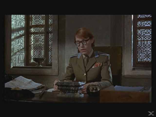 Eine Frau in Uniform sitzt an einem Schreibtisch. Sie trägt eine Brille und blickt nach unten. Fenster mit komplizierten Mustern sind im Hintergrund.