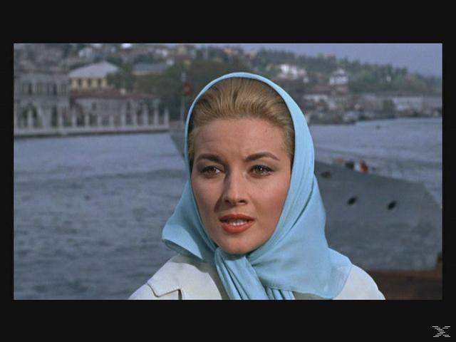 Eine Frau mit blondem Haar, die ein hellblaues Kopftuch trägt, blickt den Betrachter an. Ein Schiff und Gebäude befinden sich im Hintergrund.