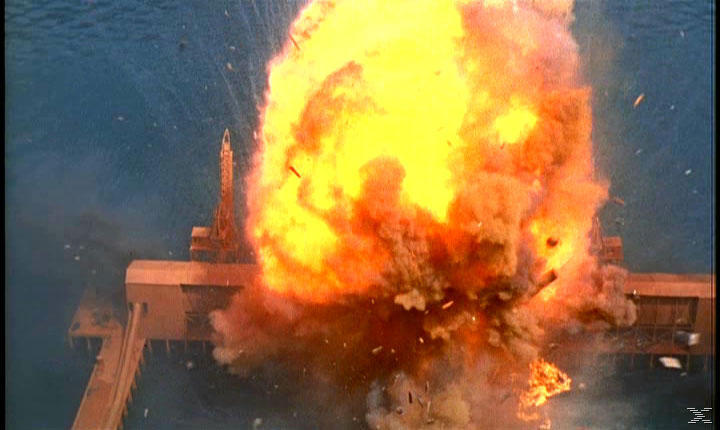 Eine Explosion bricht über einer Ölplattform aus. Flammen und Trümmer erfüllen die Luft vor blauem Meereshintergrund.