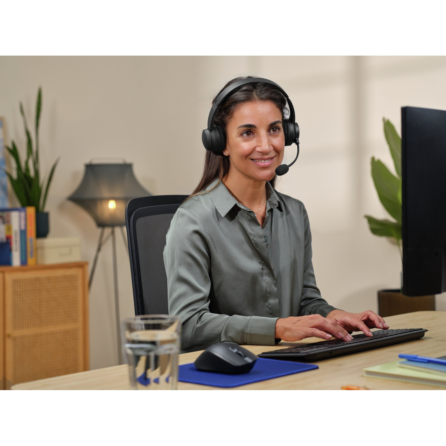 Una donna con le cuffie digita su una tastiera, sorridendo a uno schermo del computer. È in un ambiente d'ufficio.
