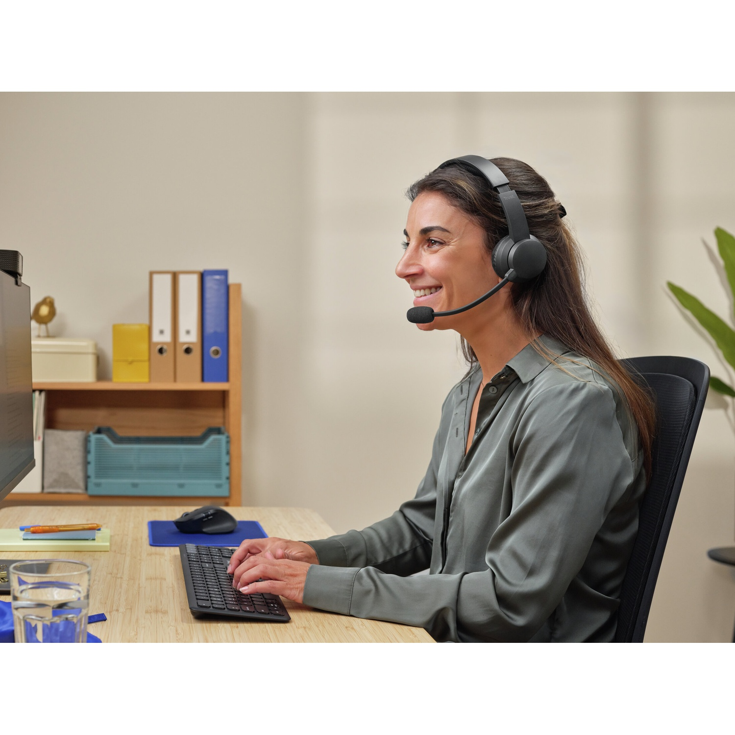 Una donna con le cuffie digita su una tastiera a una scrivania, con uno schermo di computer e forniture per ufficio sullo sfondo.