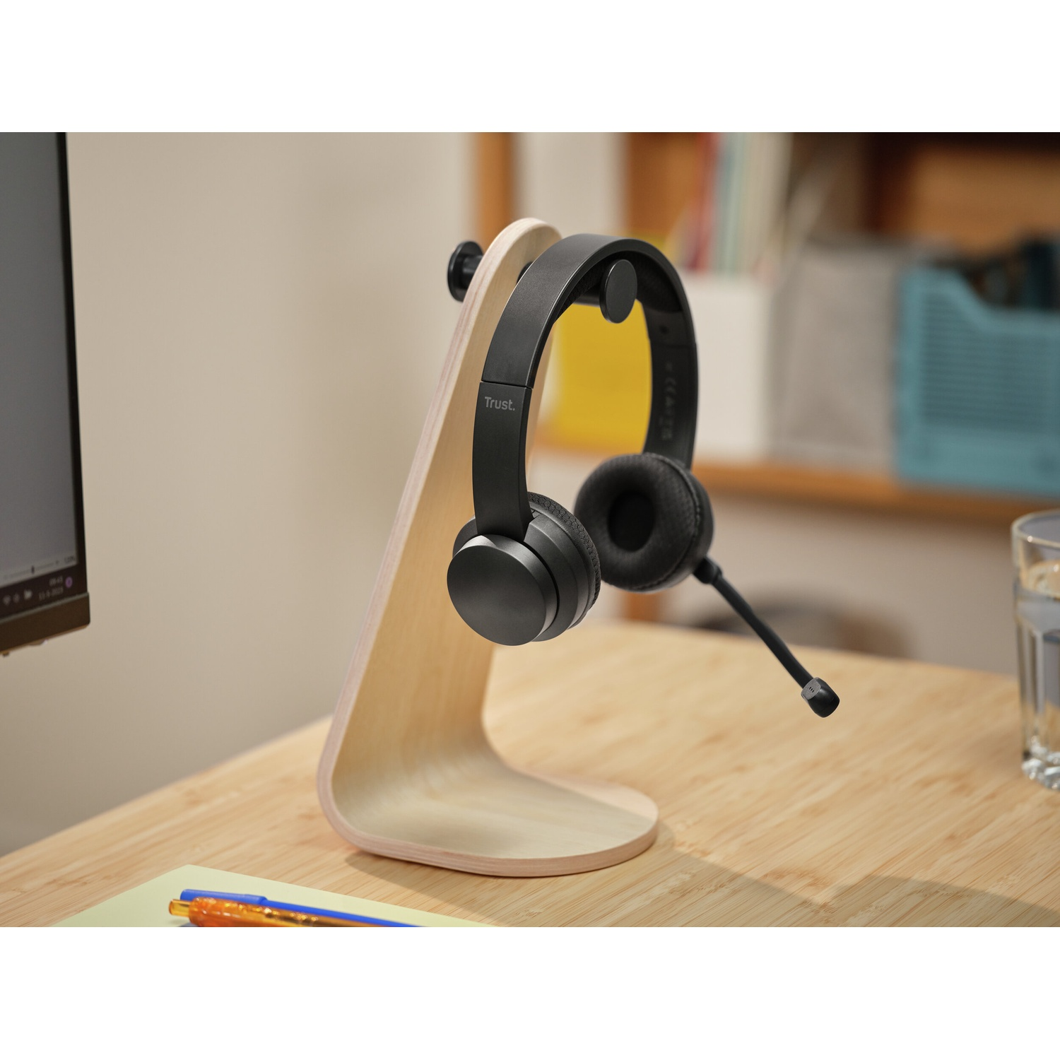 Cuffie nere su supporto in legno. Scrivania con penne, bicchiere d'acqua e computer. Lo sfondo include una mensola e un muro.