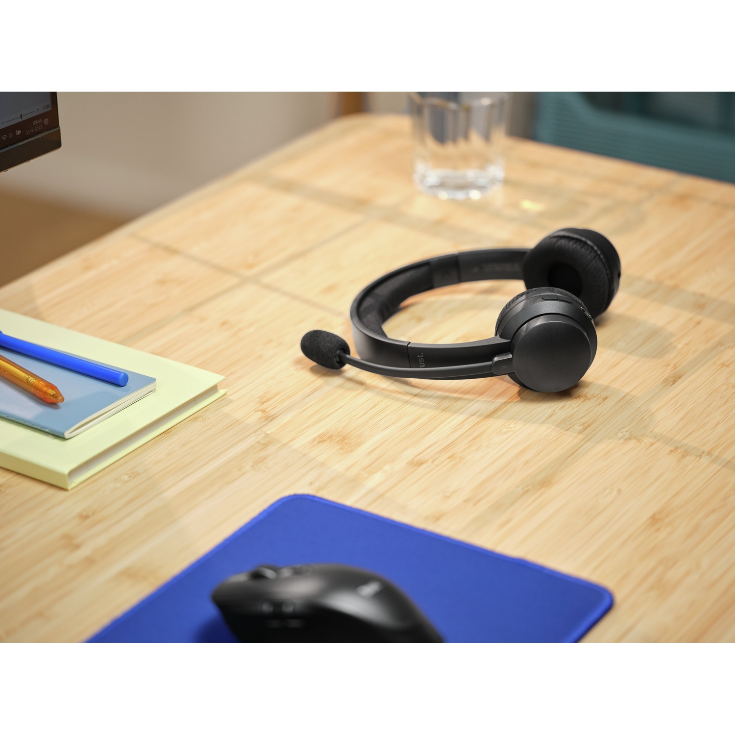 Cuffie, penne, quaderno, mouse e bicchiere su scrivania in legno. Tappetino blu per mouse. Cuffie nere con microfono. Bicchiere d'acqua.
