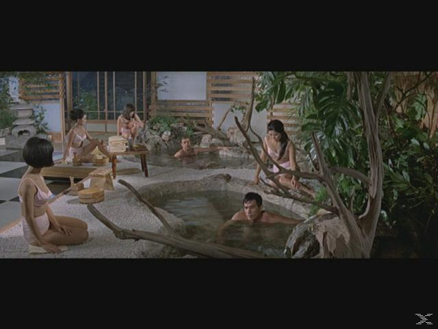 Mehrere Personen in einem Spa. Frauen tragen rosa Badeanzüge. Männer sind im Wasser. Grünes Grün umgibt die Pools mit dekorativen Steinen.